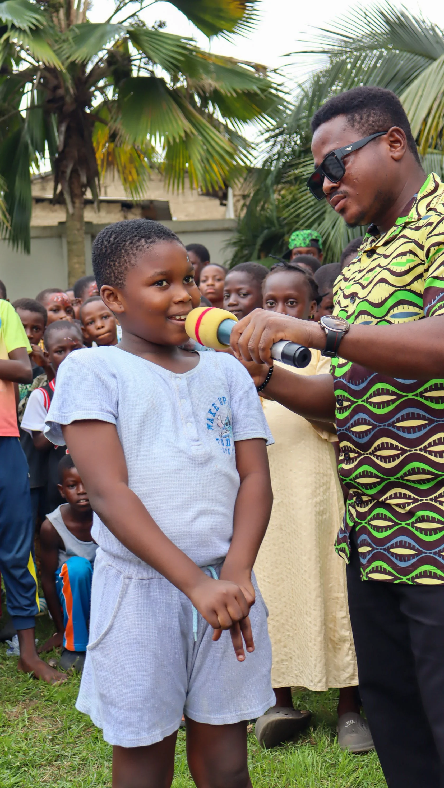 A young girl speaks into a microphone held by a man wearing sunglasses during an outdoor event, with a group of children and a palm tree in the background.