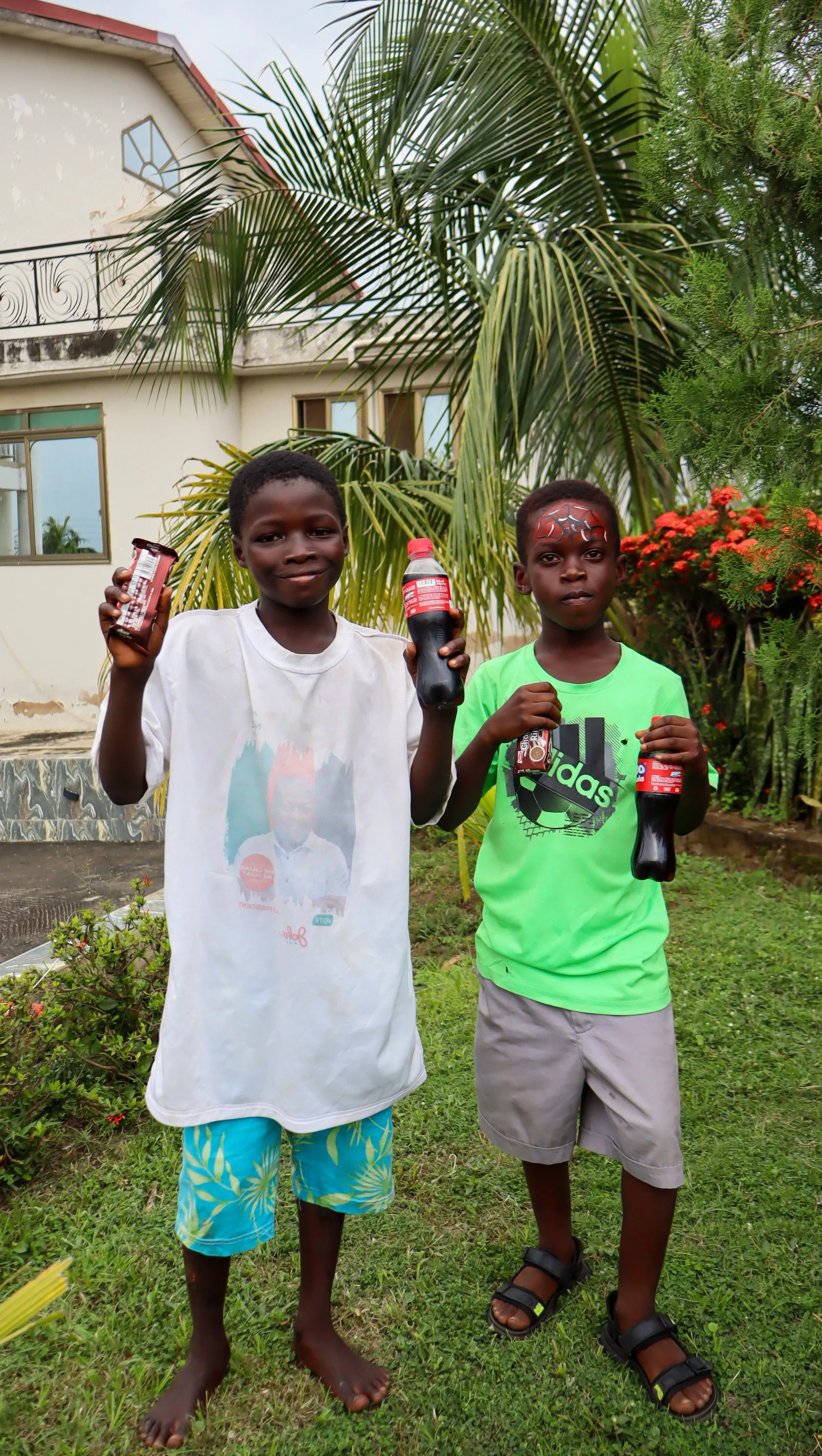 Two young boys standing outside on a grassy area, holding bottles of Coca-Cola, with tropical plants and a building in the background.