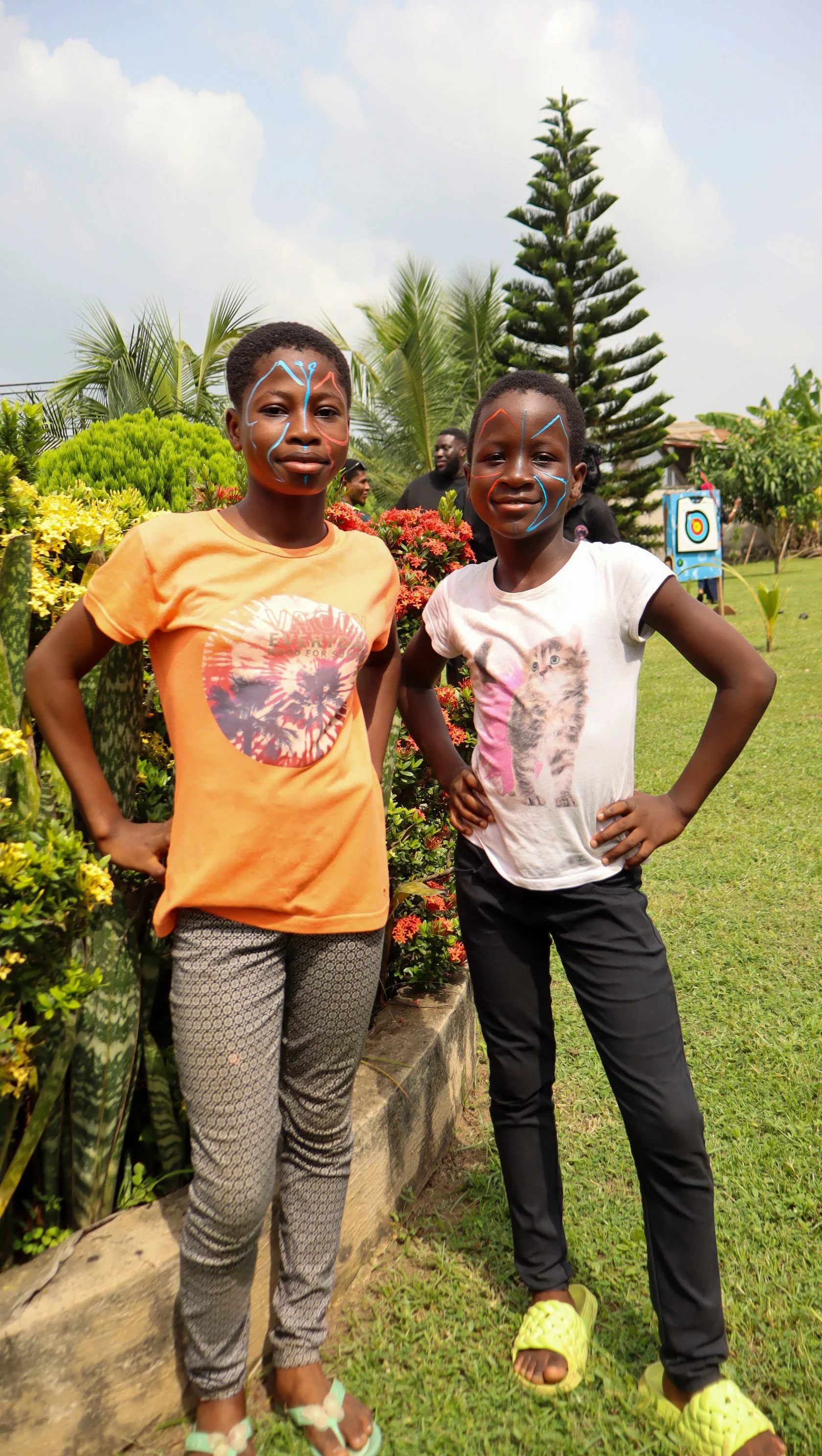Two young girls standing outdoors in a garden, smiling at the camera. They have face paint designs, and are wearing casual clothes and slippers.