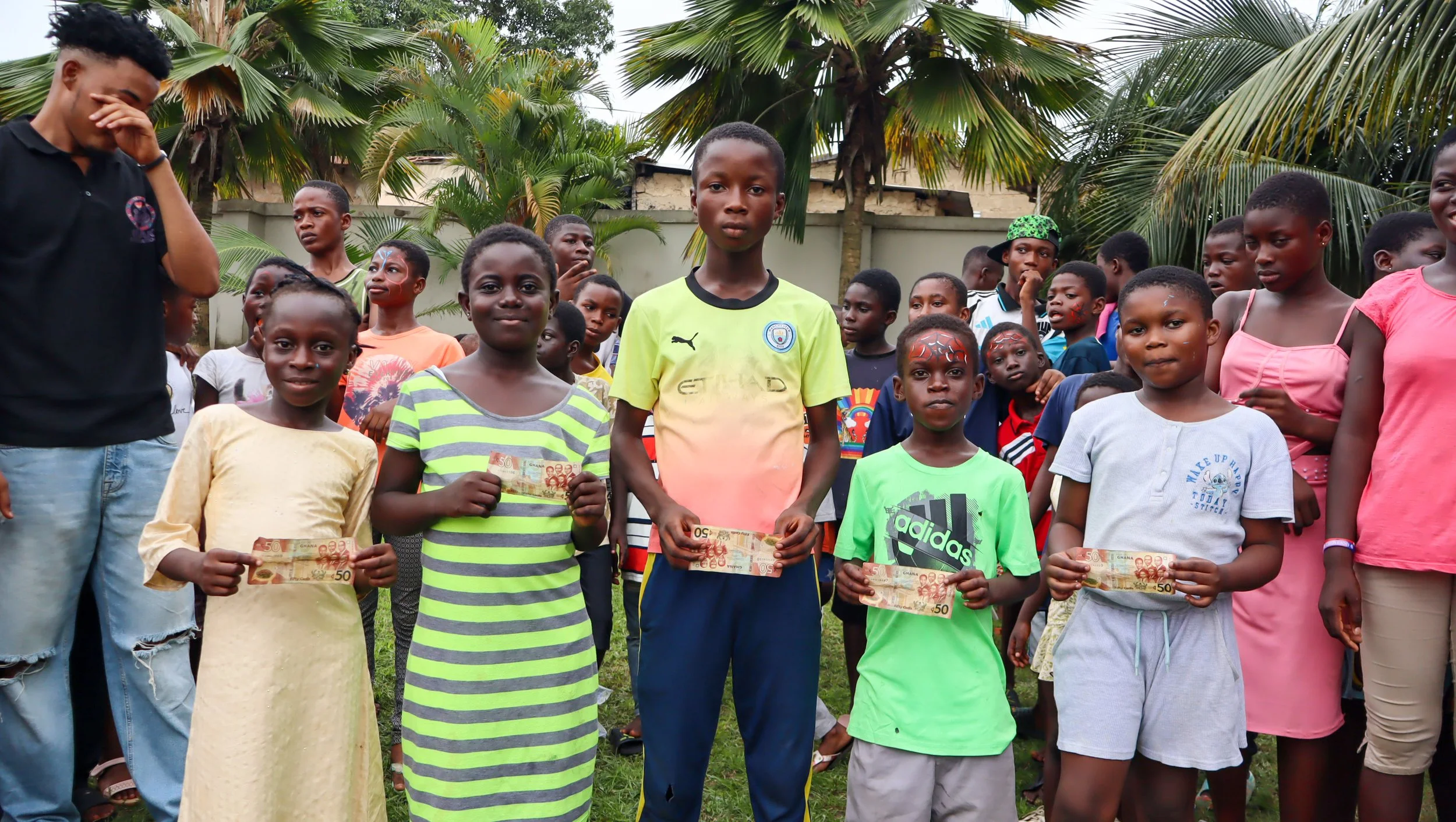 Group of children standing outdoors, some holding money, with palm trees in the background, during daytime.