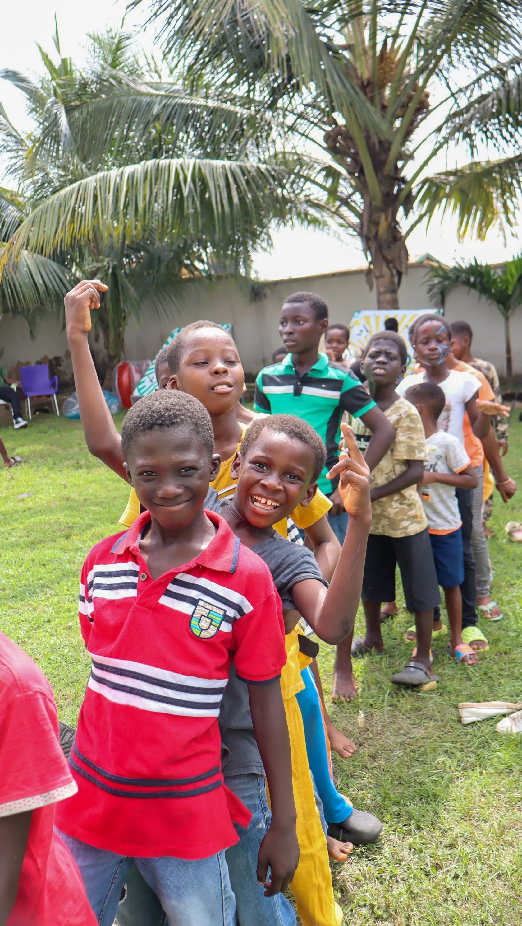 Happy children standing in a line outdoors in a grassy area, with palm trees in the background, smiling and looking at the camera.