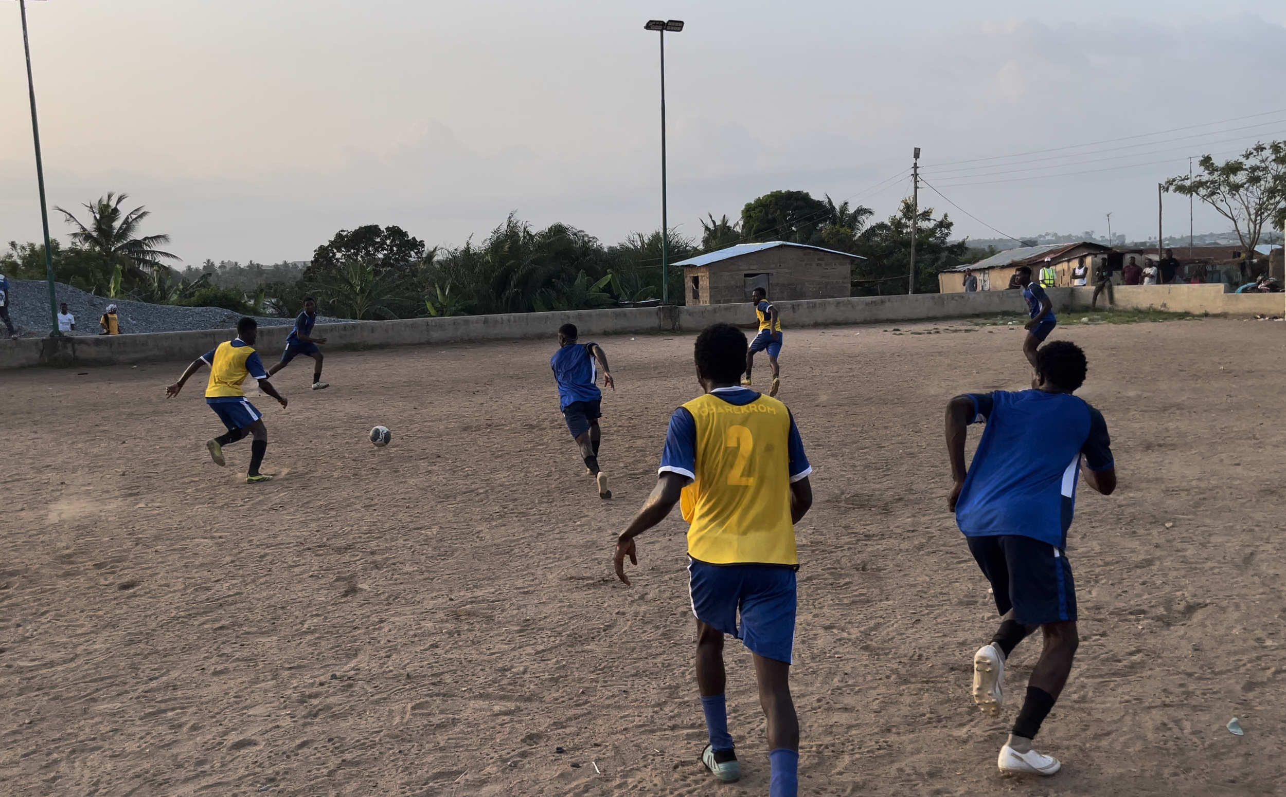 Young boys playing soccer on a dirt field with houses and trees in the background.