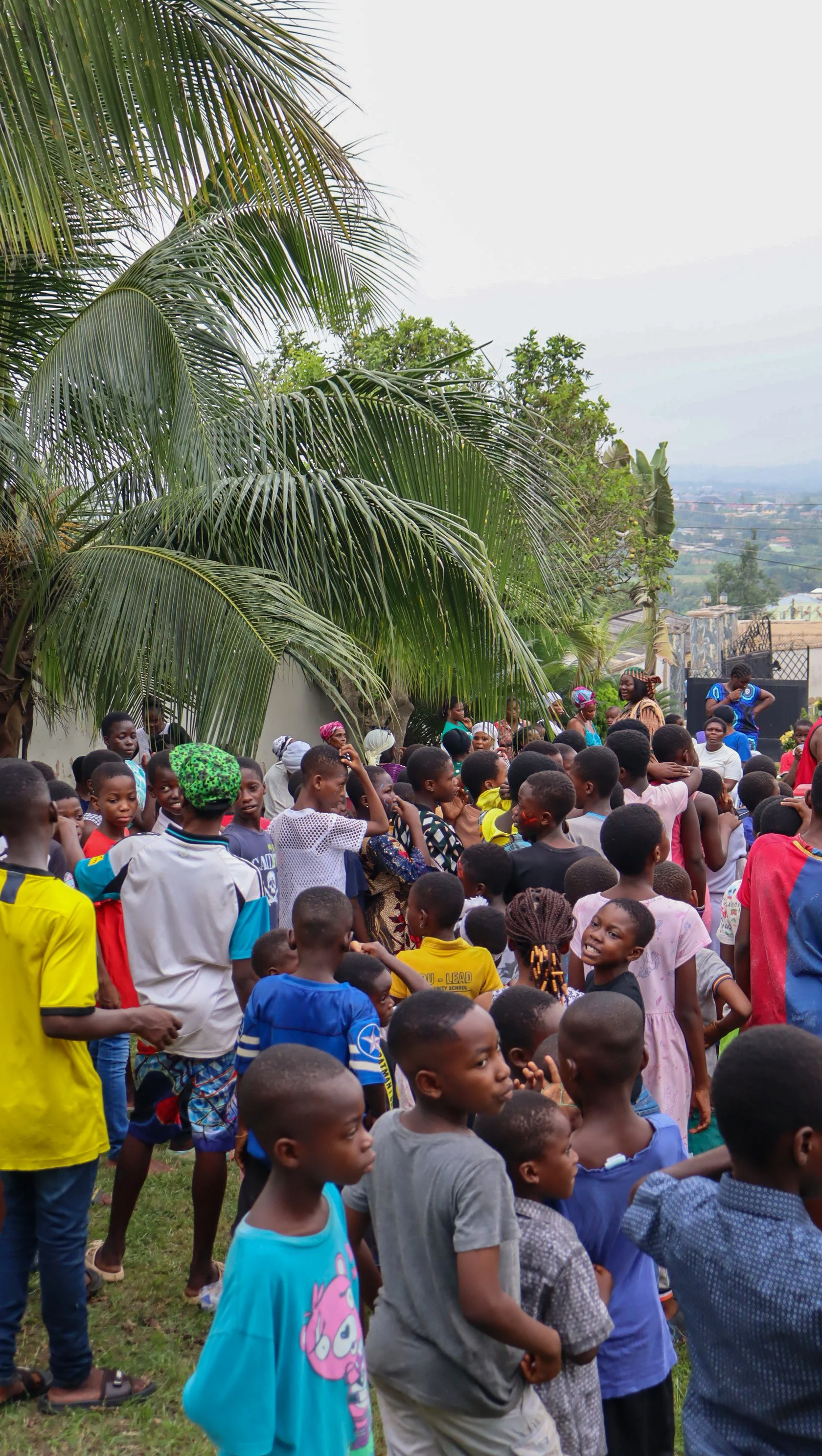 A group of children gathered outdoors under palm trees, some standing and some sitting, with buildings and a cloudy sky in the background.