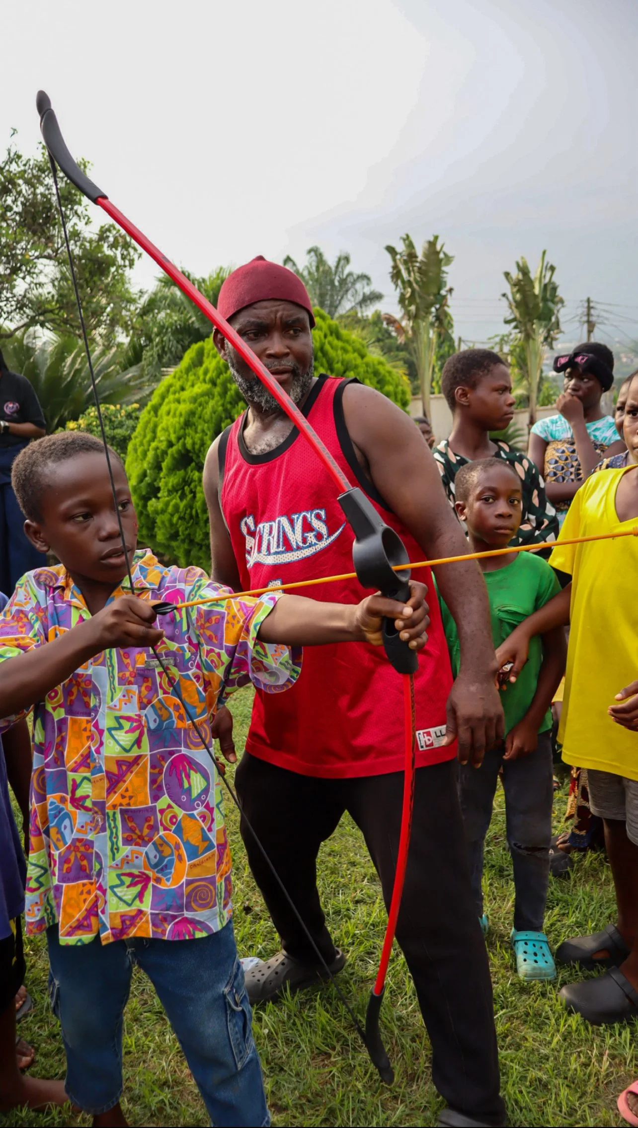 A man in a red Lakers basketball jersey and maroon beanie teaches children archery outdoors, with lush greenery and trees in the background.