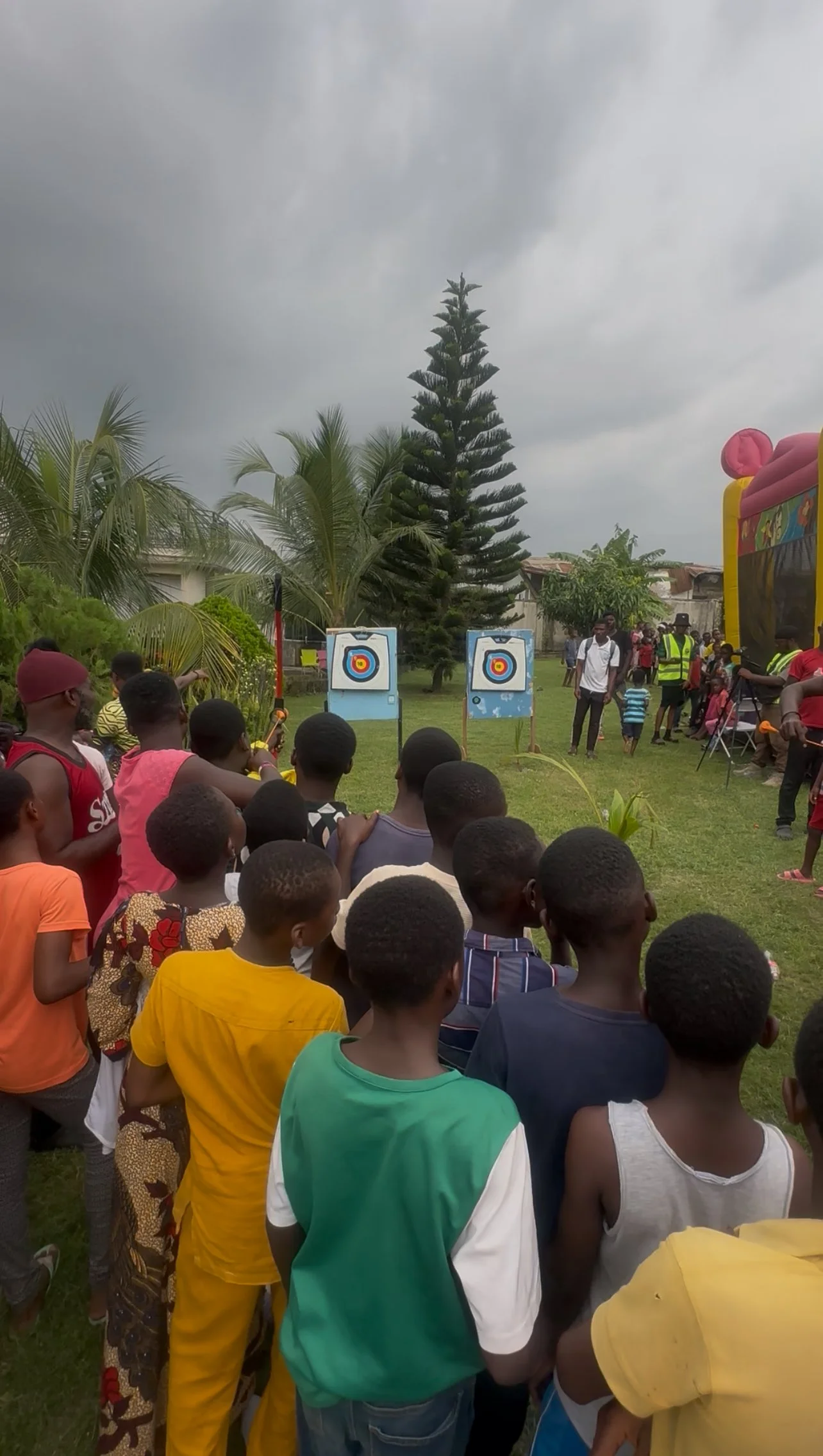 A crowd of children and adults gathered outdoors on a cloudy day, watching an archery game with two targets set up on stands. There are trees and a colorful inflatable structure in the background.
