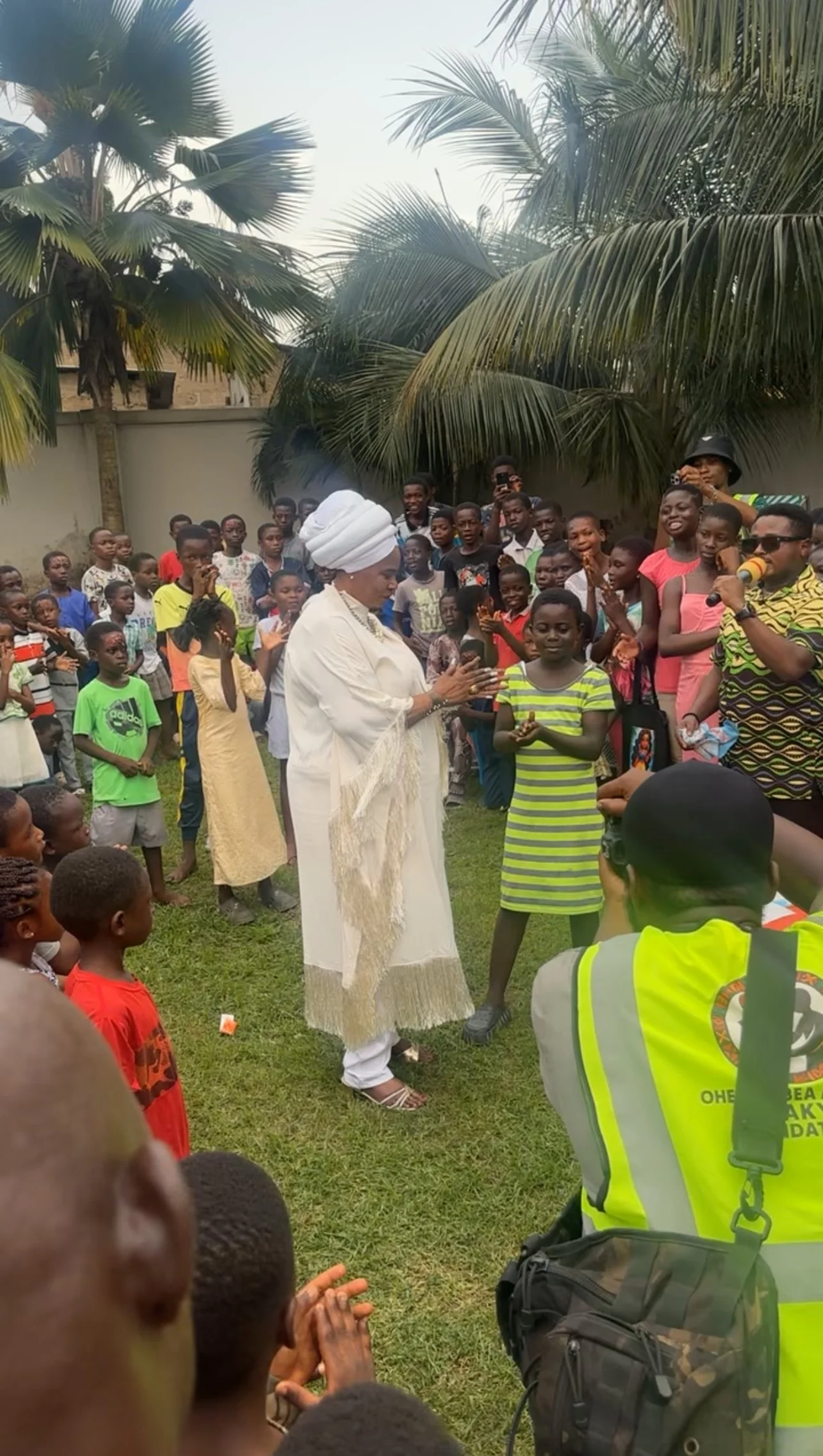 A woman dressed in white traditional attire and a head wrap stands in the center of a crowd of children and adults, clapping and smiling outdoors with palm trees in the background.