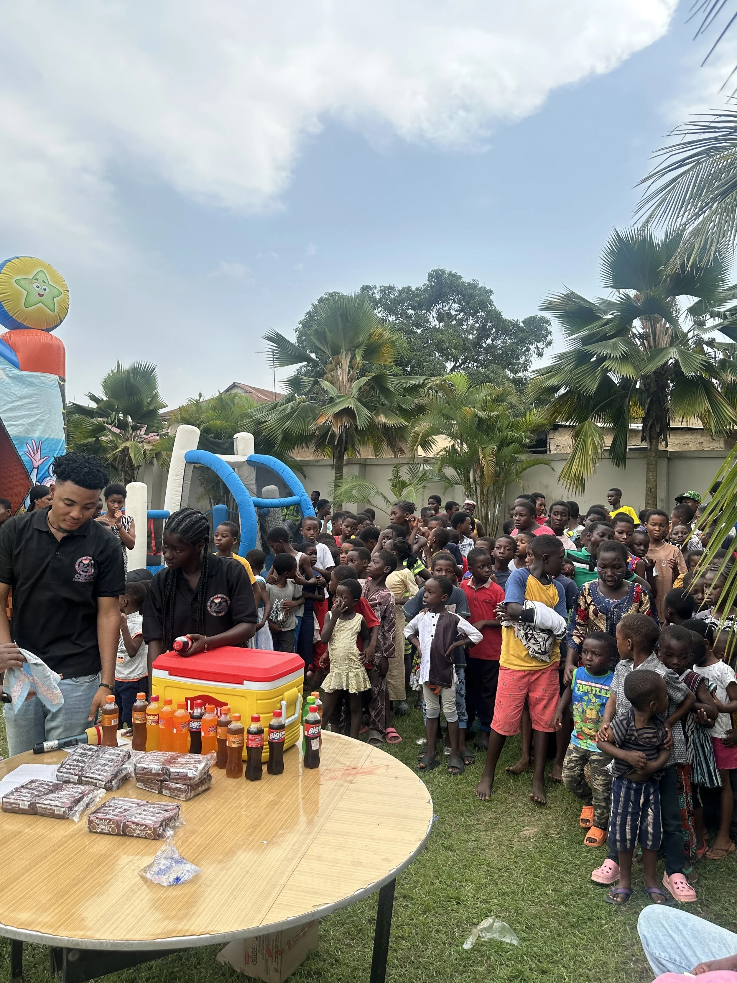 Children and adults gather outdoors at a party with a bounce house and tables of food and drinks.