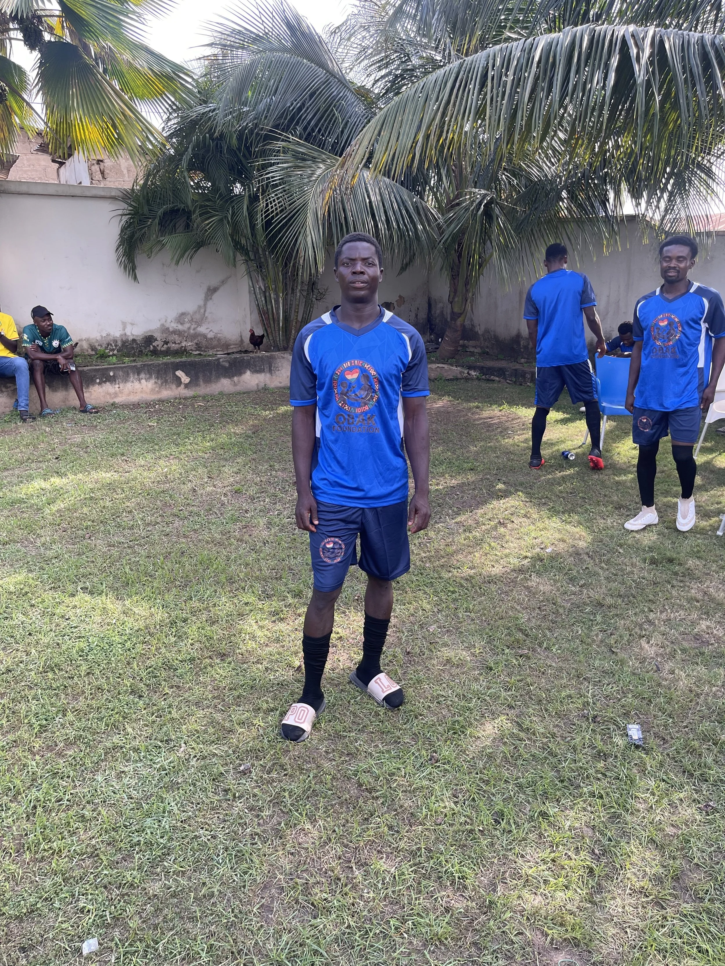 A young man in a blue soccer uniform standing on a grass field, with other players and people in the background under a large palm tree.