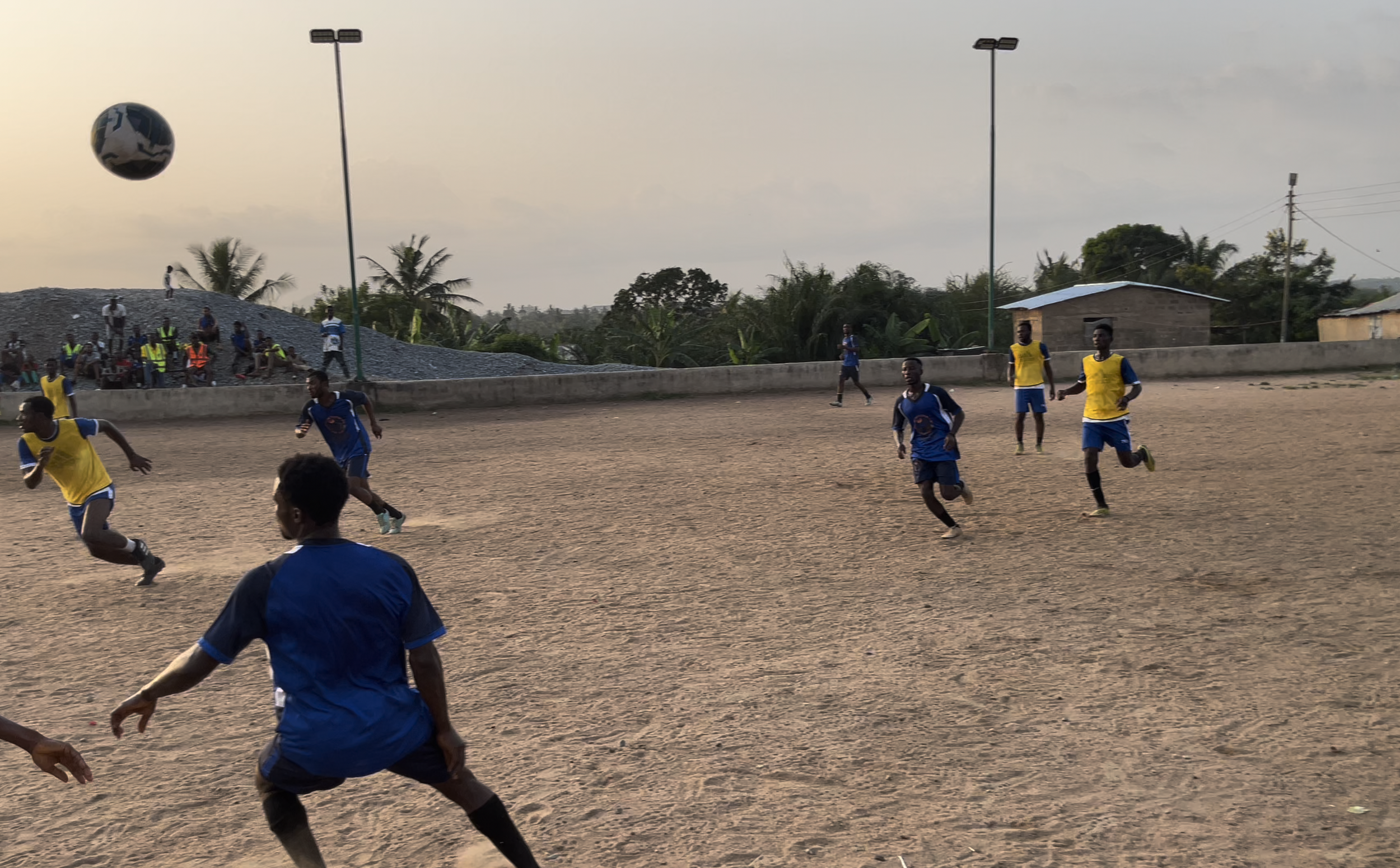 Children playing soccer on a dirt field, some wearing yellow bibs and others in blue uniforms, with spectators sitting on a mound in the background, under a cloudy sky.