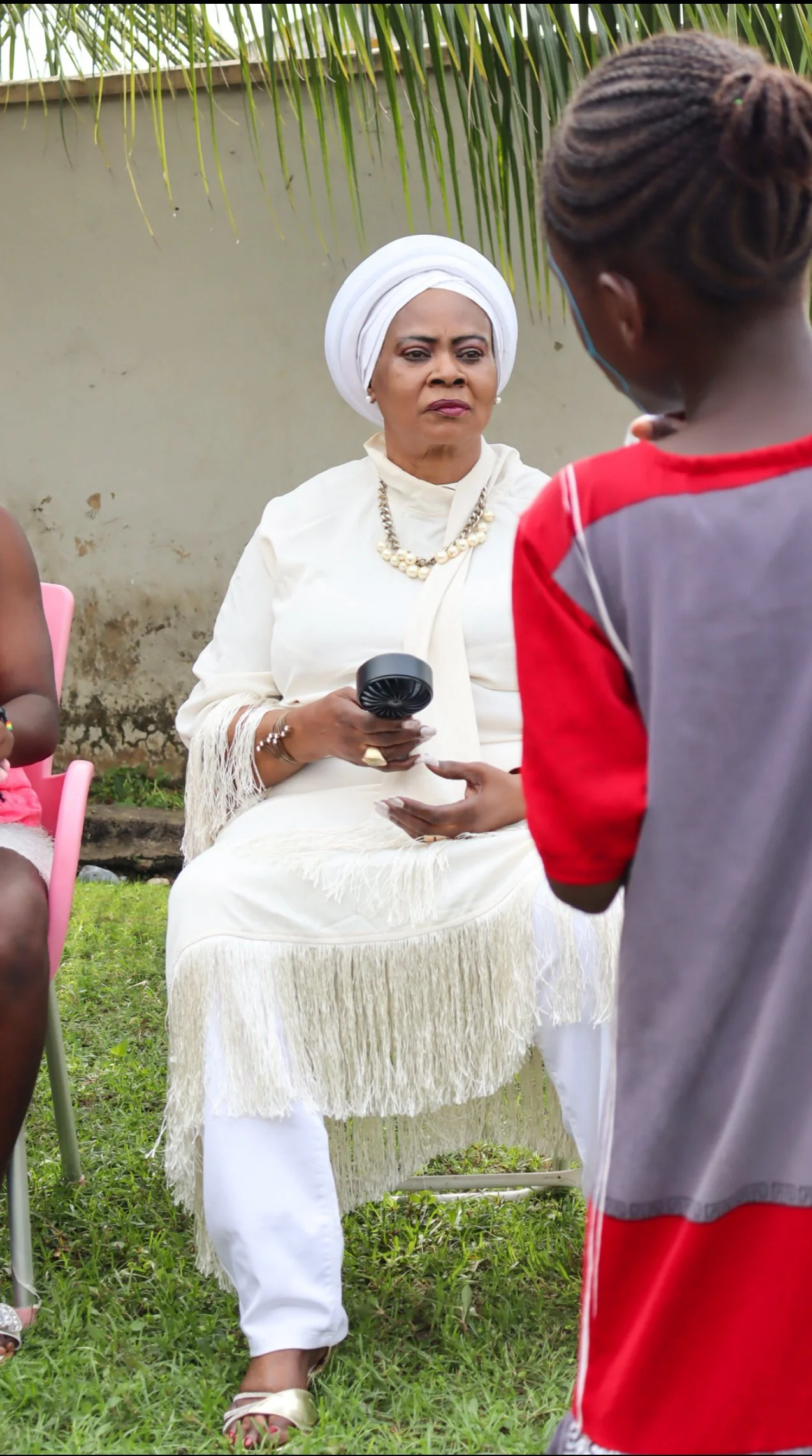 An elderly woman dressed in white with a turban and jewelry sits outside on grass, holding a small black fan, while a young boy in a red and grey shirt stands in front of her, talking to her. There are other people partially visible, and a palm tree 