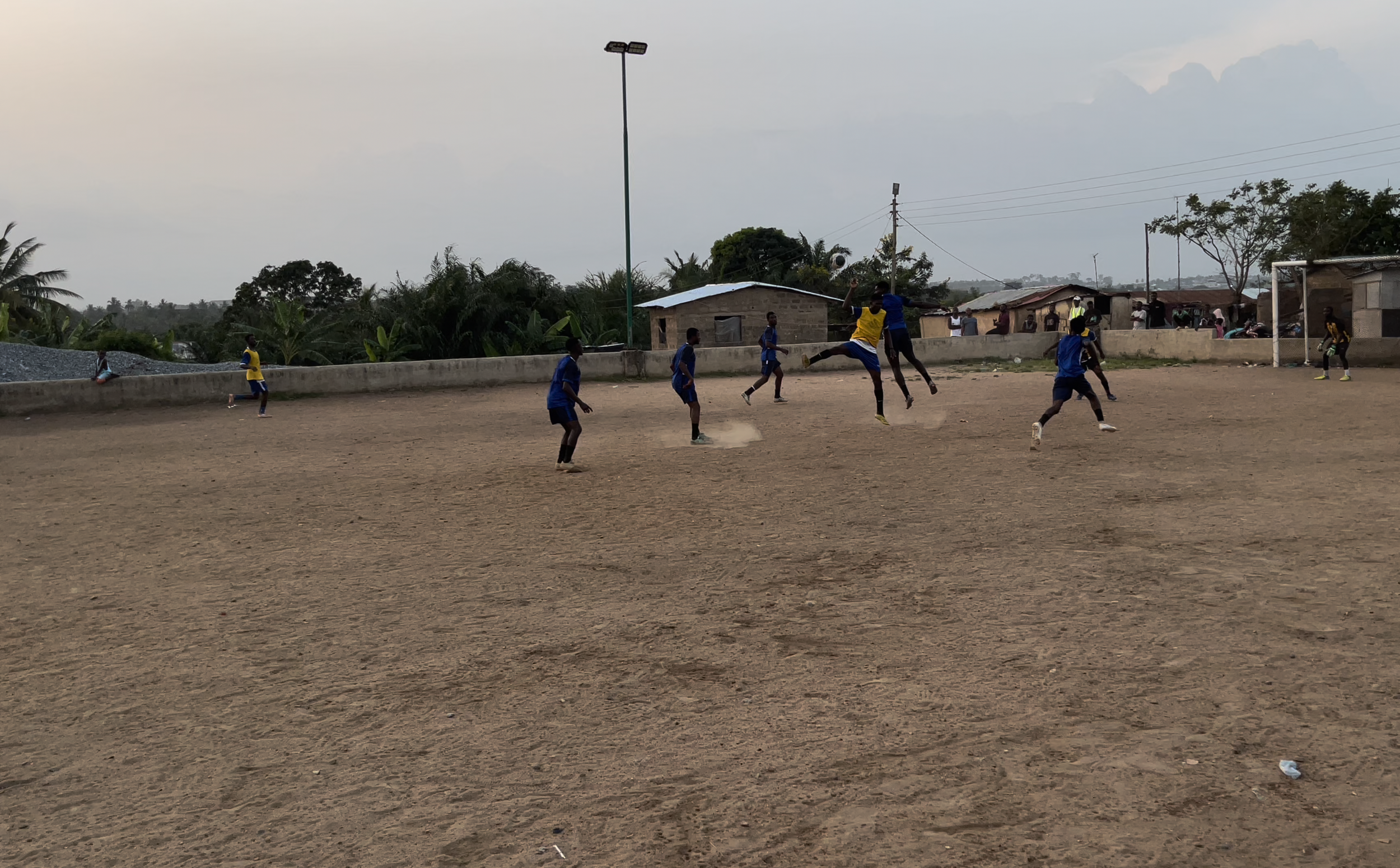 Young boys playing soccer on a dirt field in a rural area, with houses, trees, and a cloudy sky in the background.