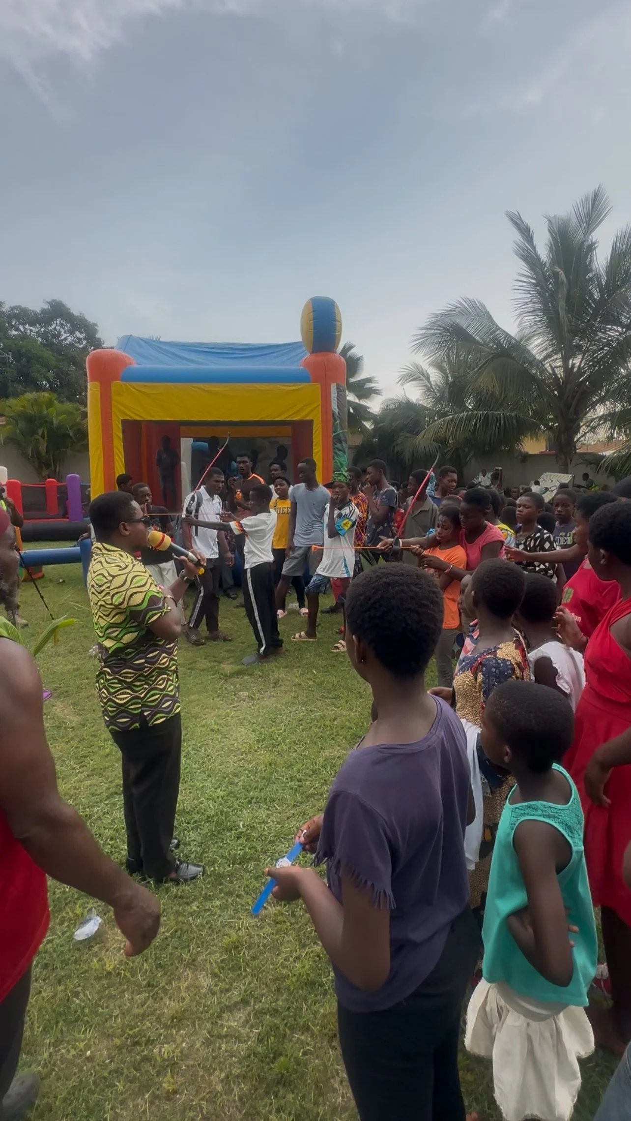 Children and adults at a birthday party beating each other with blow-up batons in a circle outside, with an inflatable bounce house in the background.