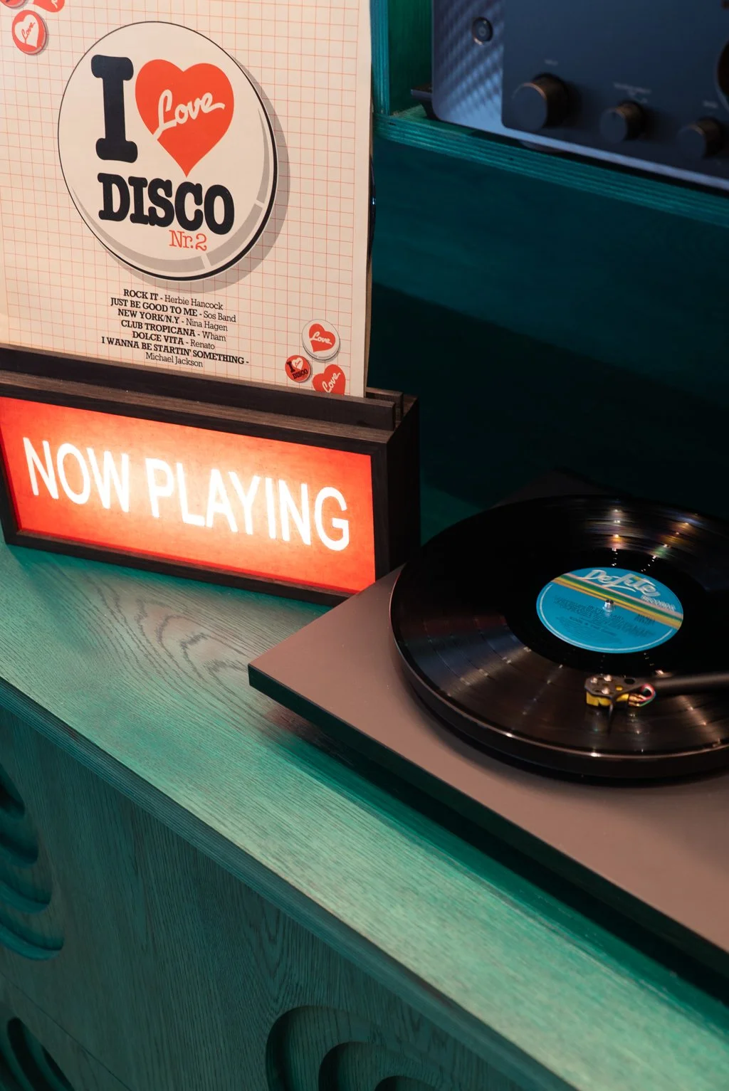 A record player on a light green wooden table with a lit sign that reads "NOW PLAYING." Next to it, a stylized sign features the album cover for "I Love Disco" with a list of disco songs. The setup is in a dimly lit space with teal and dark blue walls.