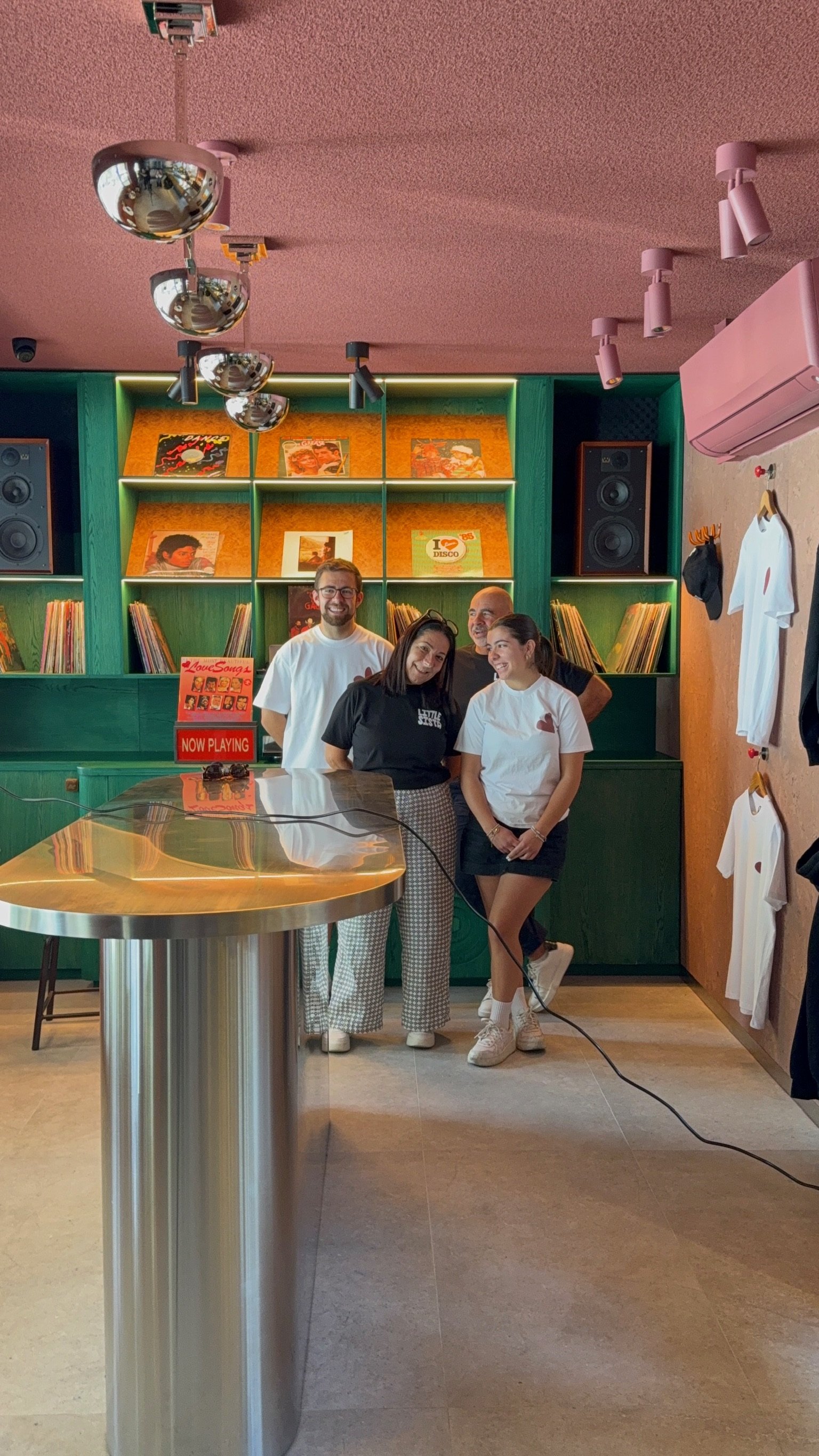 Group of four people standing inside a record store, smiling for the camera. The store has green shelves with vinyl records and album covers in the background, and a table with a sign that reads 'Now Playing' in the foreground.