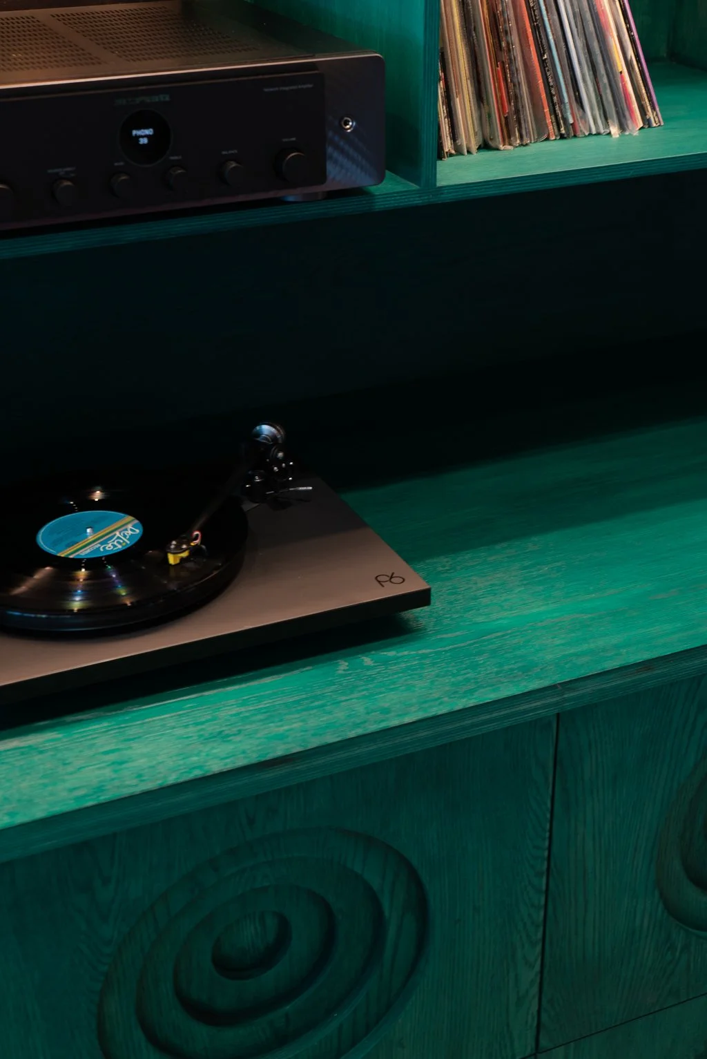 A turntable playing a vinyl record on a green wooden cabinet, with a black audio receiver and a collection of vinyl records on a shelf above.
