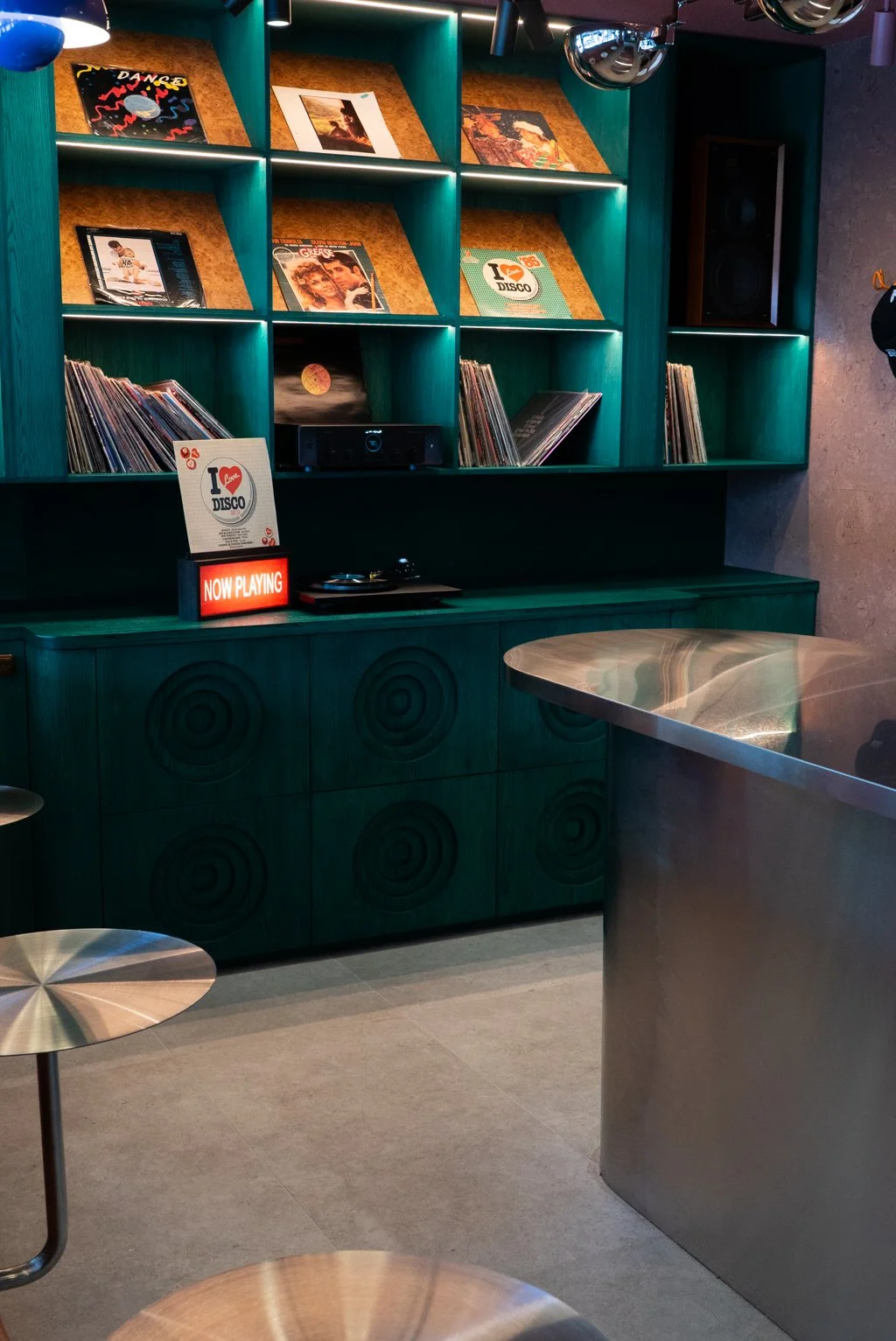 A teal and wooden vinyl record shelf with posters, records, and a small turntable, in a modern indoor space with a metal counter and stools.