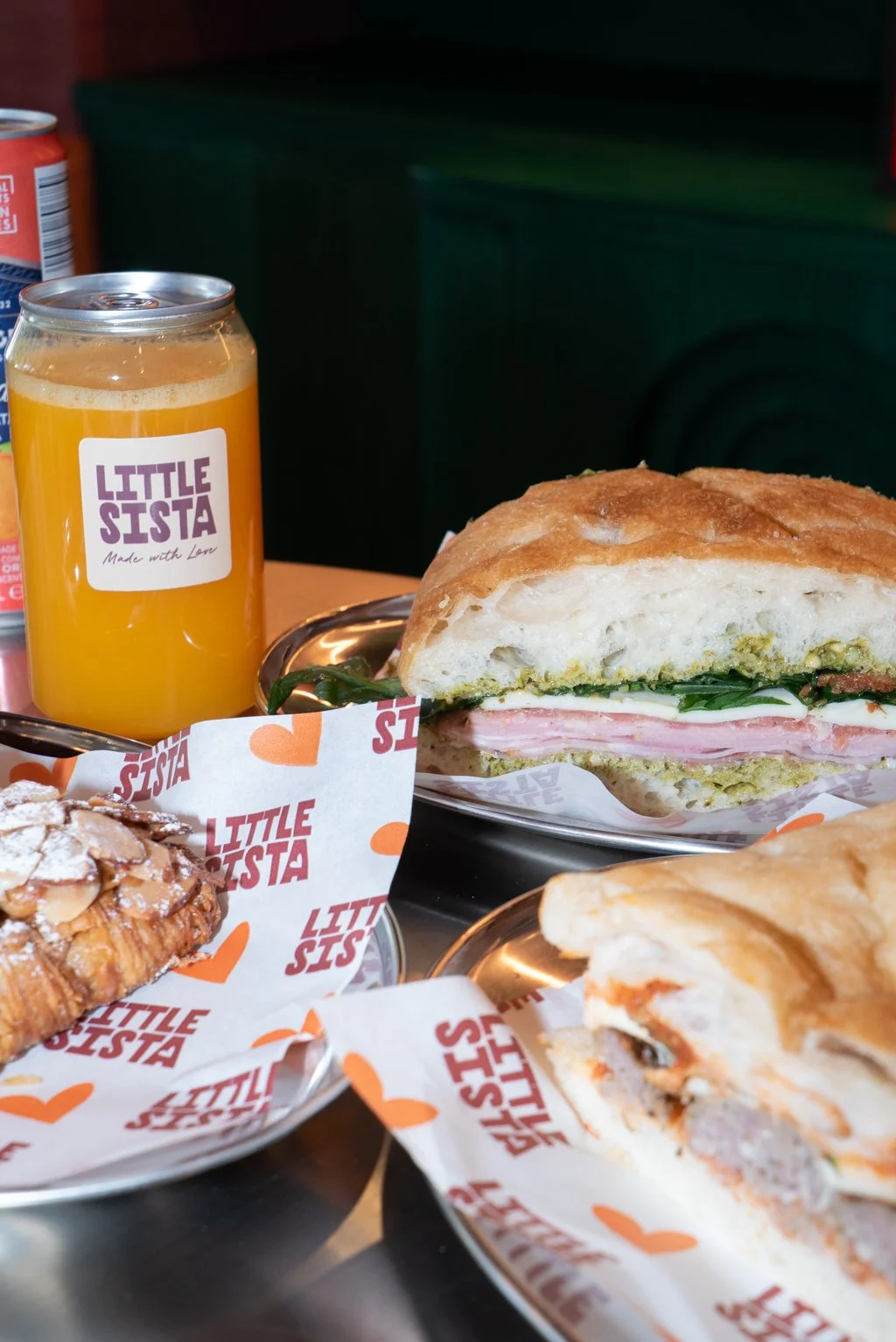 Sandwich with ham, spinach, and sauce on a toasted bun, a glass of orange juice with a "Little Sister" label, a pastry with powdered sugar and almonds, and a sandwich with prosciutto and cheese on a paper-lined tray.