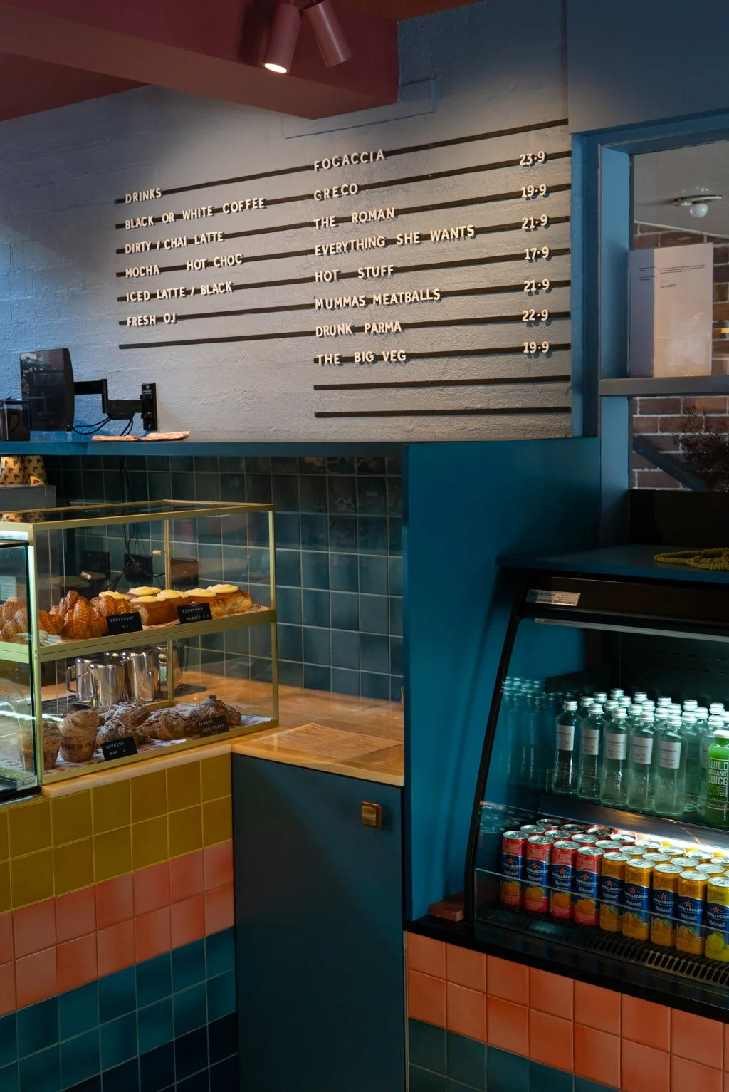 Menu board listing various food and drink options with prices, and a display case with pastries, coffee, and beverages at a cafe.