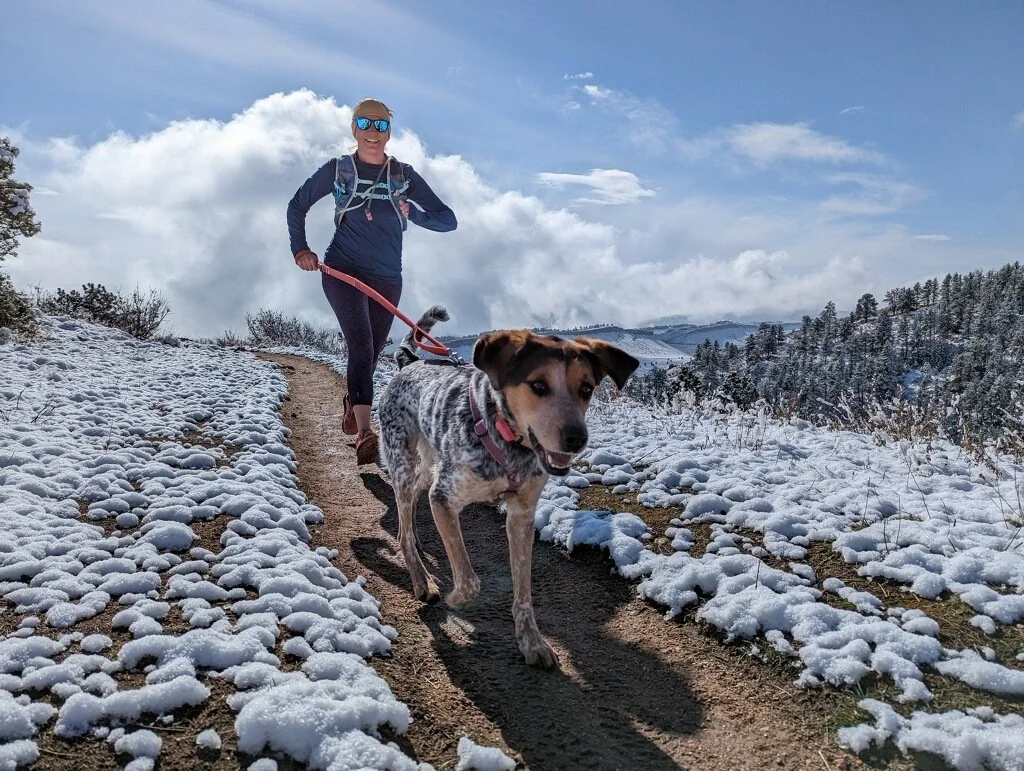 A woman walking her dog on a snow-covered trail during daytime in a winter landscape with snow on the ground, trees, and hills in the distance.