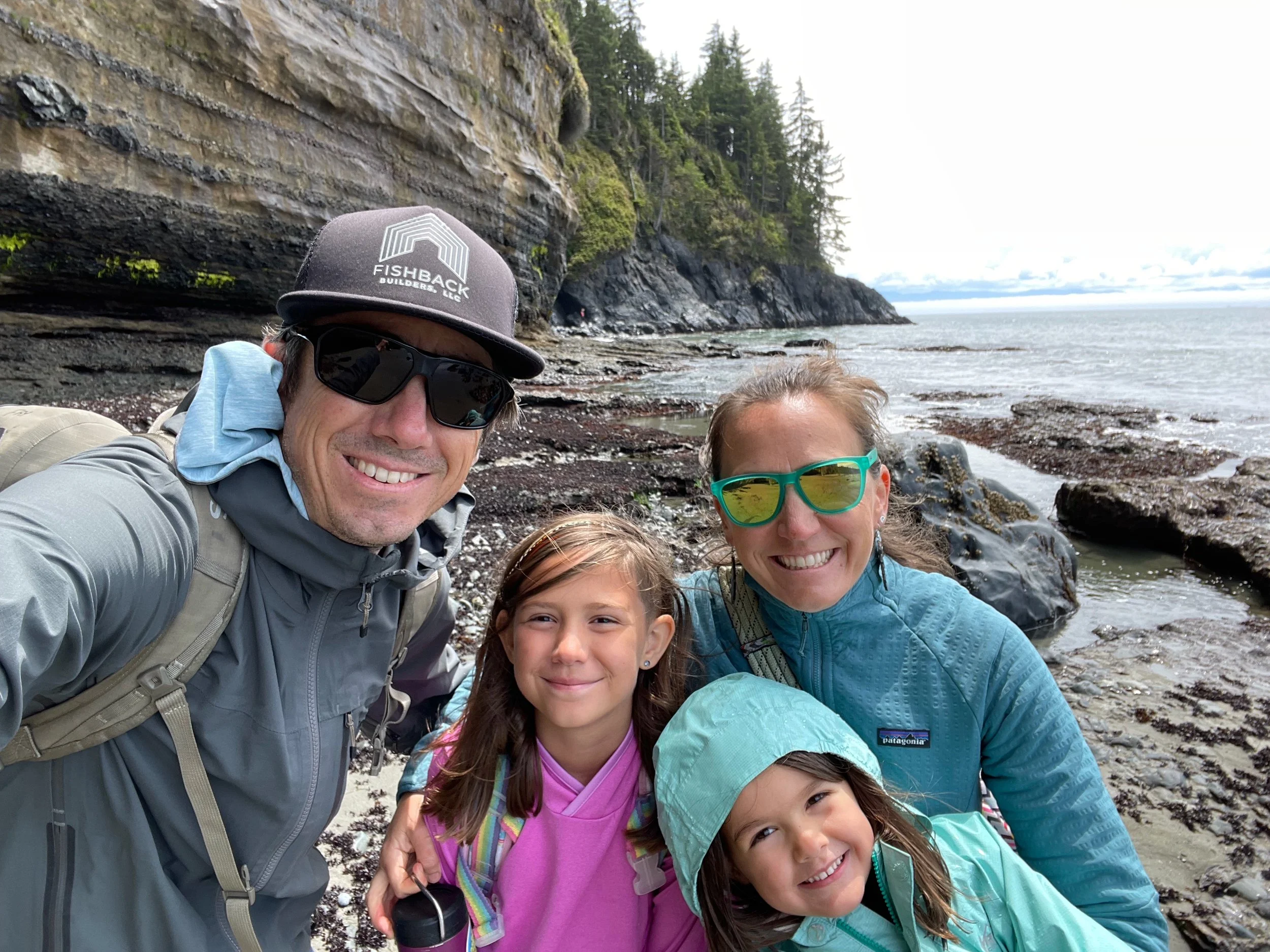 Family of four taking a selfie on a rocky beach with cliffs and ocean in the background. The man is wearing a gray jacket, sunglasses, and a black hat with a logo, and the woman is wearing a blue jacket and sunglasses. The girls are smiling, one in a pink jacket with a backpack, and the other in a teal jacket with a hood.