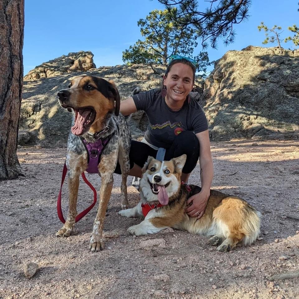 A woman smiling and crouching outdoors with two dogs on a dirt ground, surrounded by rocks and trees, under a clear blue sky.