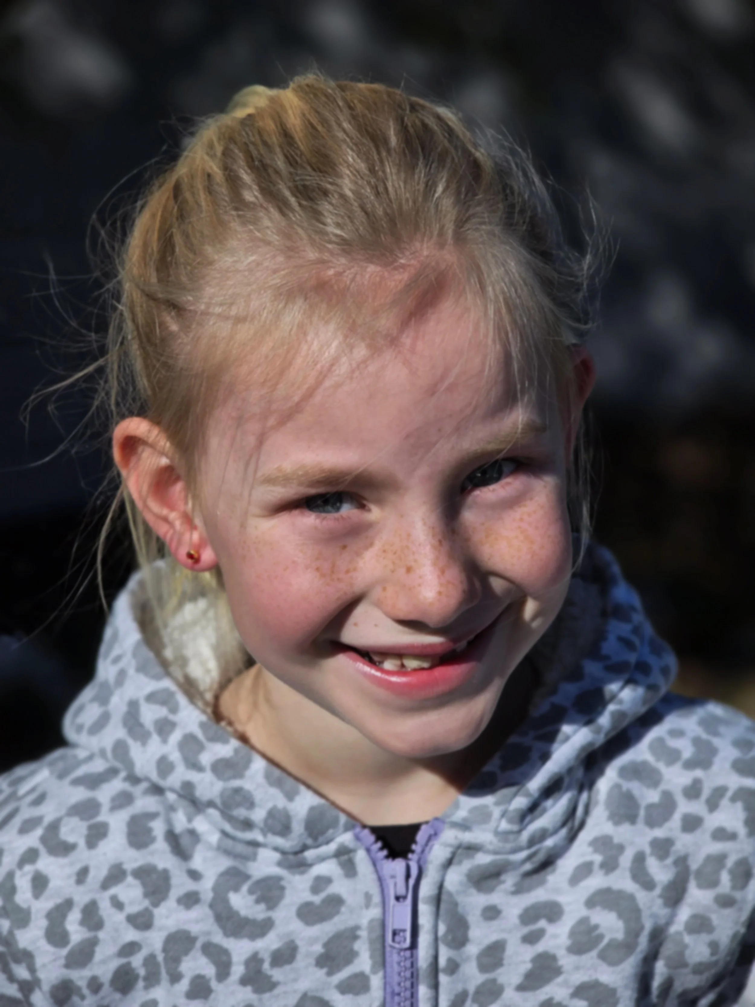 Close-up of a smiling young girl with blonde hair, freckles, blue eyes, and wearing a gray patterned hoodie.