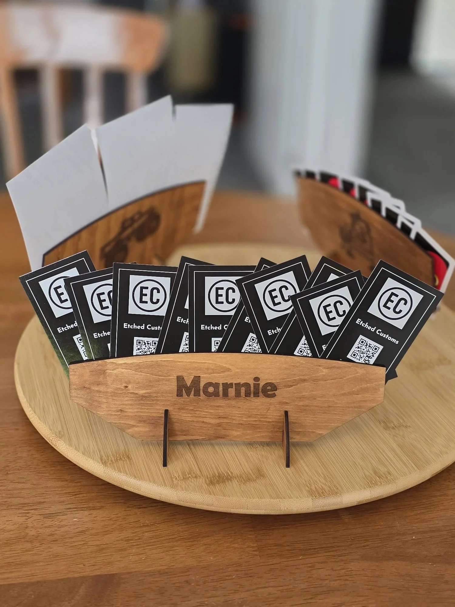 A wooden boat-shaped stand on a round wooden tray, holding business cards with the logo 'EC' and the text 'Etched Customs,' with the name 'Marnie' engraved on the stand.