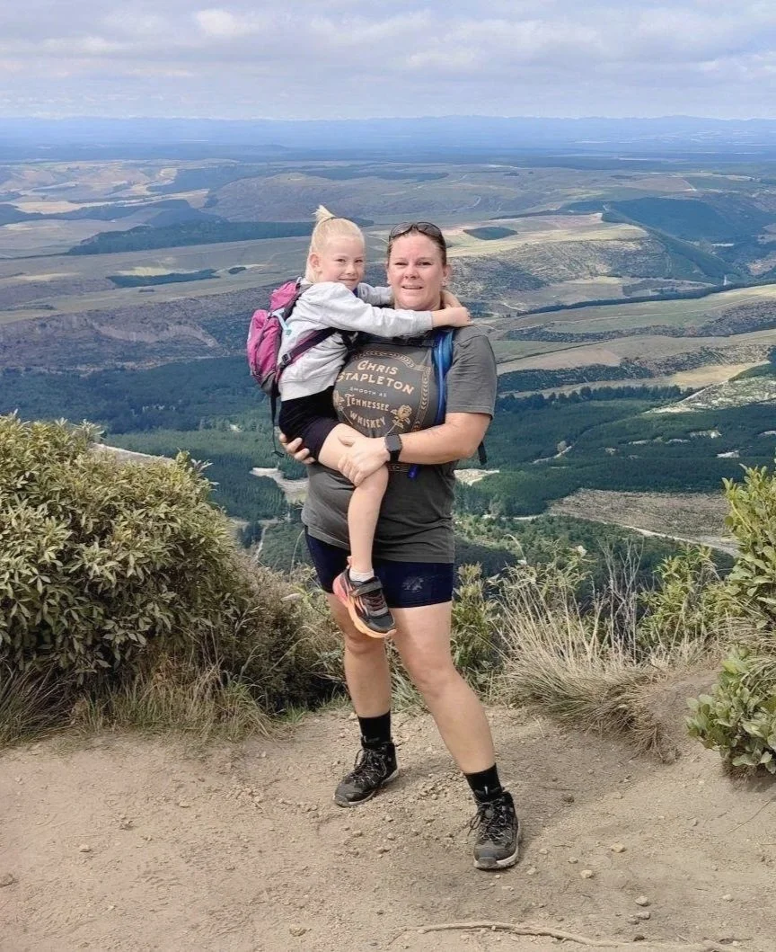 A woman holding a young girl in front of a scenic mountainous landscape with rolling hills and a cloudy sky.