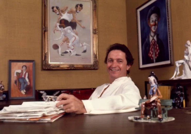 A man sitting at a desk, smiling, with various framed artworks on the wall behind him, including portraits and a painting of people playing basketball.
