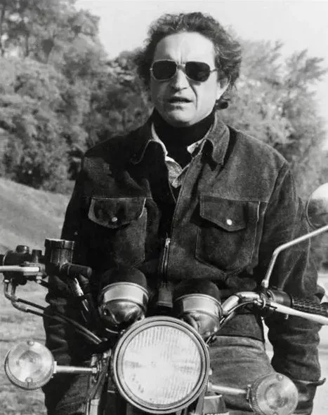 A man with curly hair wearing sunglasses and a denim jacket sitting on a motorcycle outdoors.