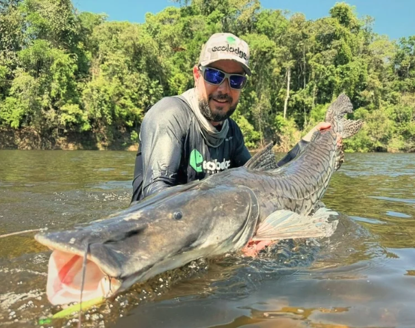 Man holding a large fish in a river with trees in the background.