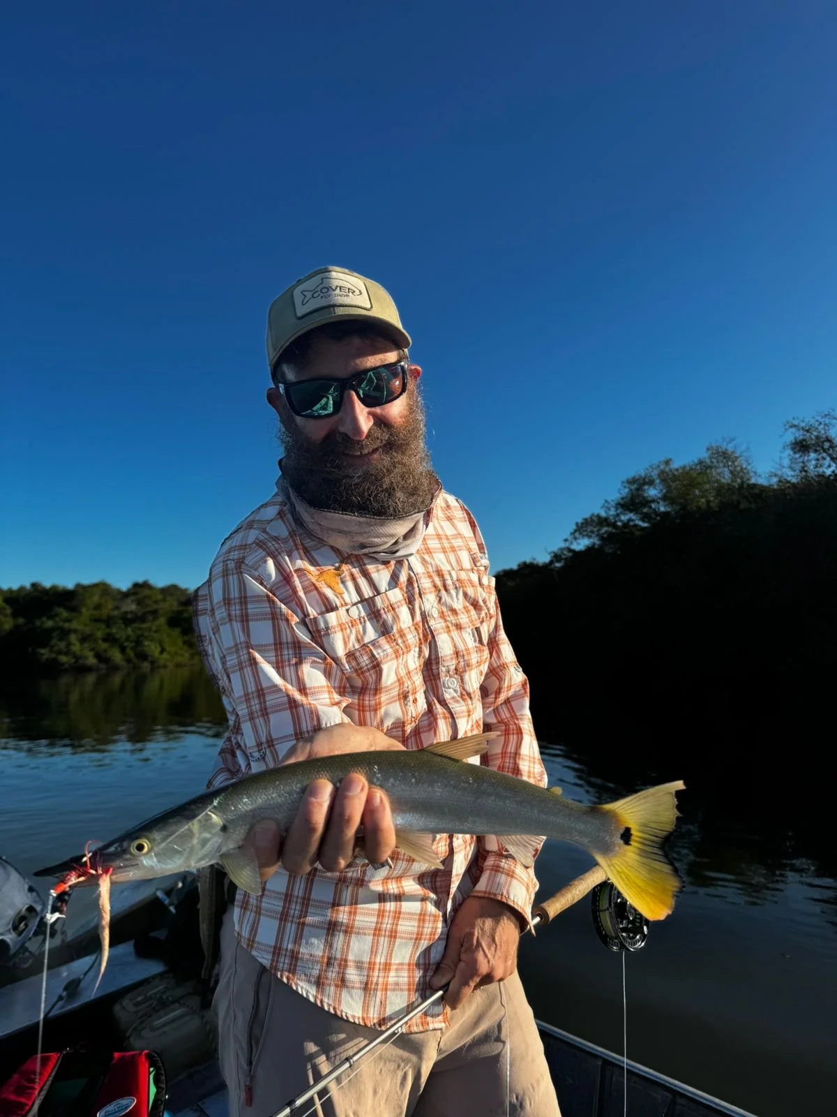 A man with a beard, sunglasses, and a cap is smiling while holding a fish he caught in a boat on a calm river with trees in the background under a clear blue sky.
