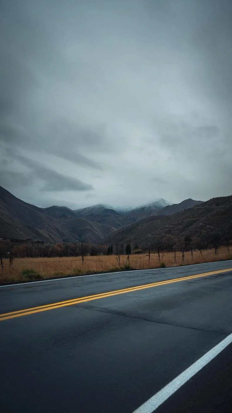 A two-lane asphalt road with yellow dividing lines runs through a mountainous landscape under a cloudy sky. Brown grass and some trees line the roadside, and snow-capped mountains are visible in the distance.