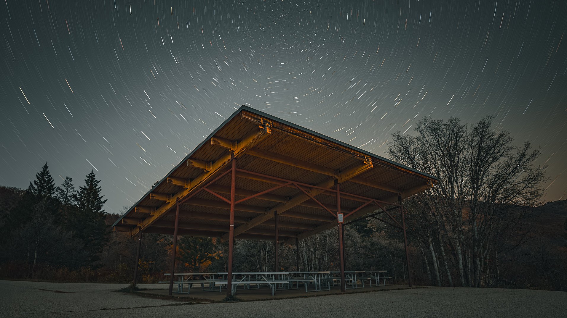 Star trails over an empty pavilion with picnic tables and trees in the background at night.