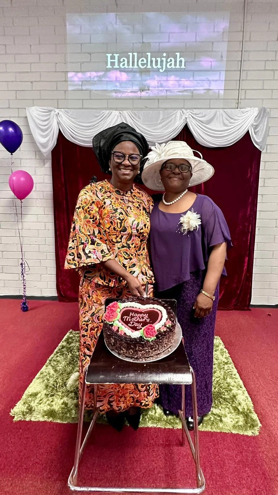 Two women in colorful and purple dresses, wearing hats and jewelry, smiling and standing behind a cake that says 'Happy Mother's Day'. They are in a decorated room with balloons, a red and white draped backdrop, and a projected 'Hallelujah' on the wa