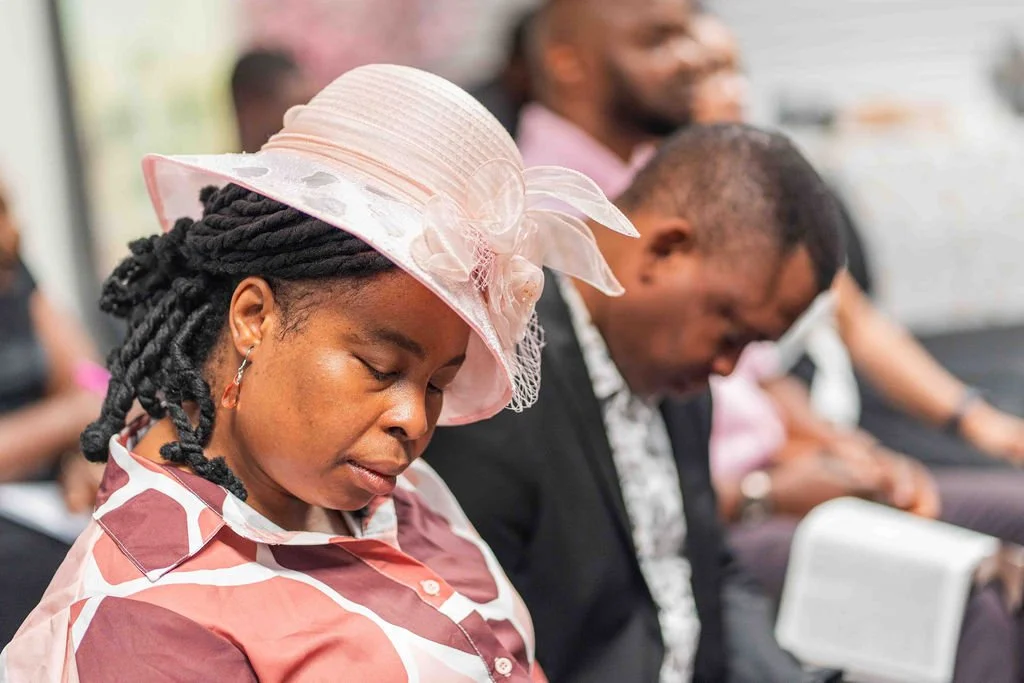 Woman wearing a pink hat and patterned shirt sitting among people at an indoor gathering