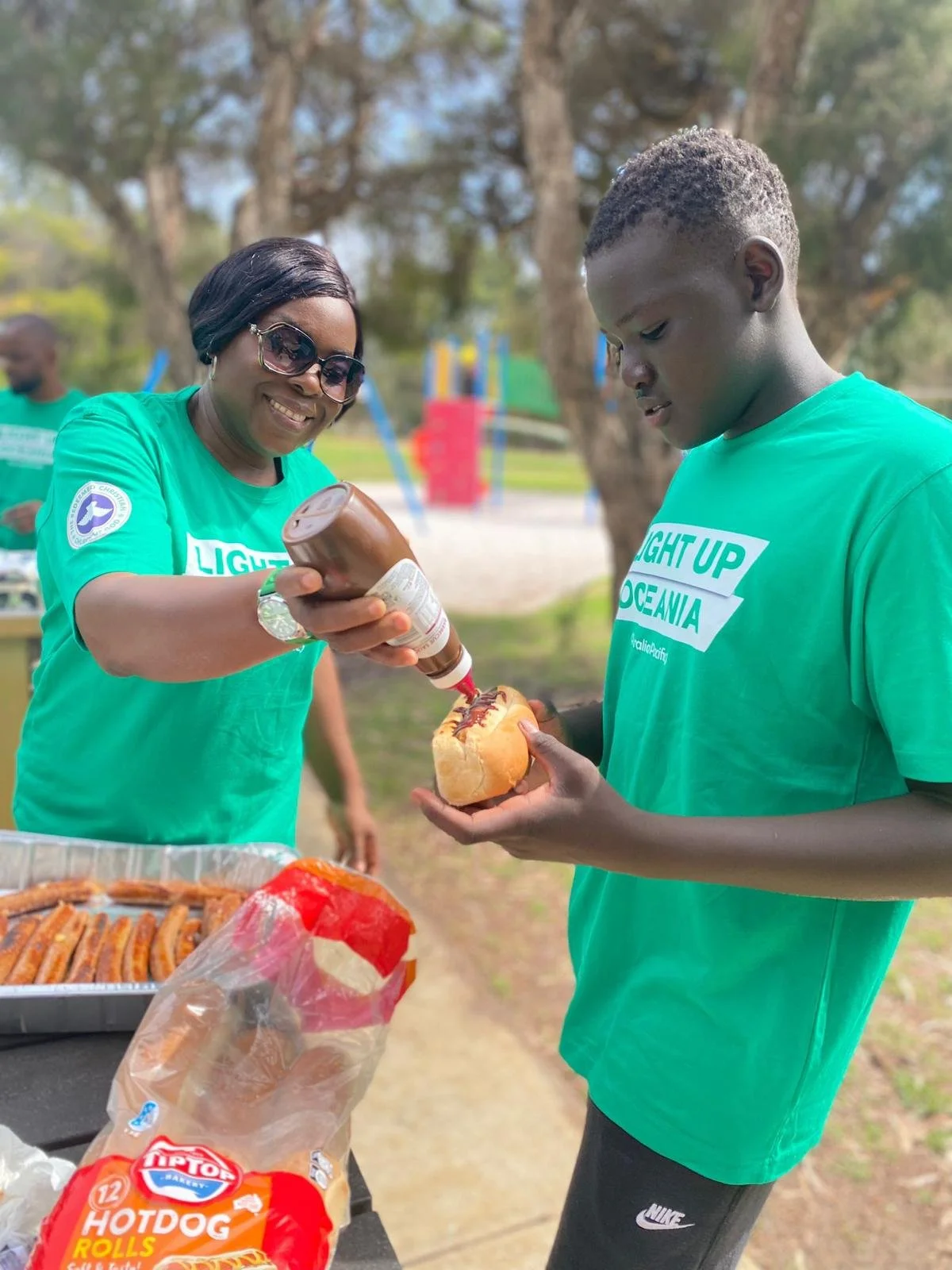 A woman and a young man at a picnic table outdoors, with the woman smiling and pouring ketchup on a hot dog held by the young man. On the table are hot dog buns and hot dogs, and the woman is wearing sunglasses and a green T-shirt.
