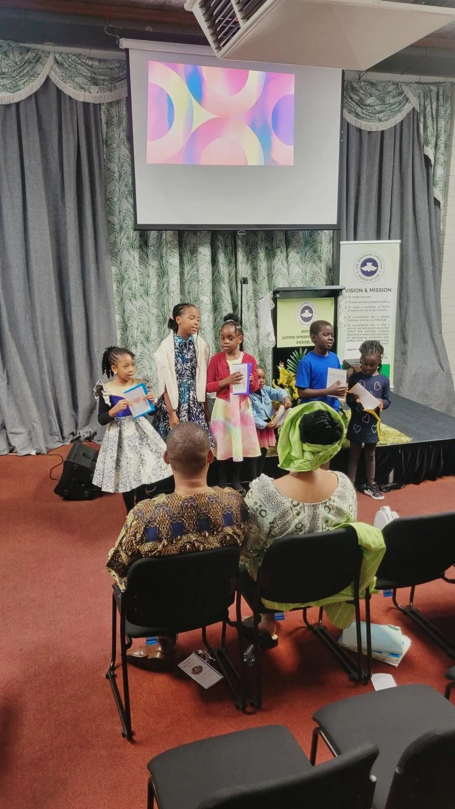 A group of children performing or presenting on stage in front of an audience in a room with gray curtains, a projector screen with colorful abstract shapes, and a banner. The audience members are seated, some wearing traditional clothing.