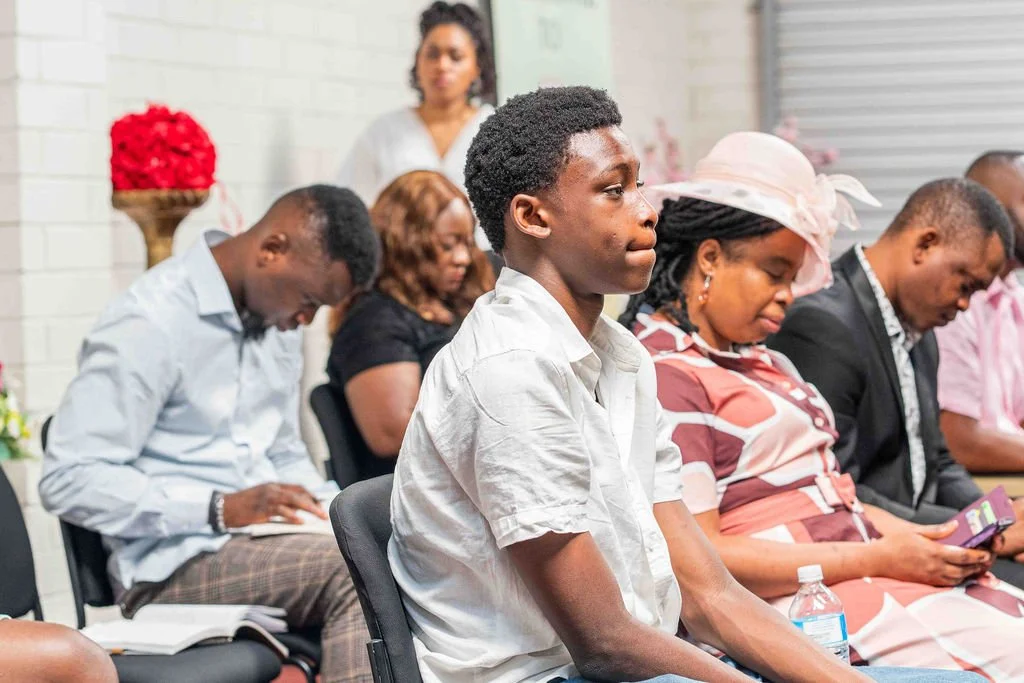 Diverse group of people seated at an indoor meeting or church gathering, with a young man in a white shirt in the foreground.