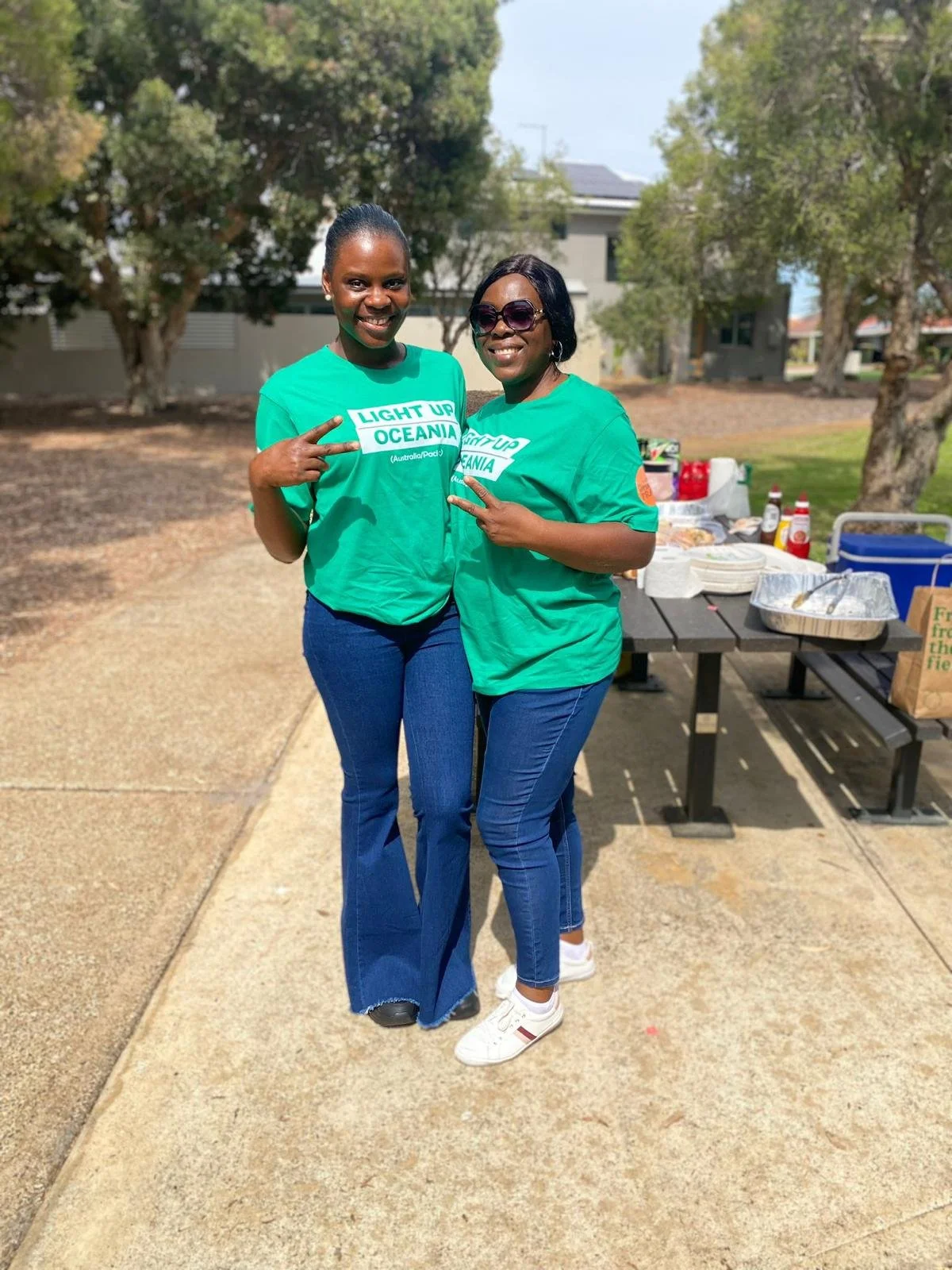 Two women smiling and posing together outdoors, wearing green T-shirts with the text "Light Up Oceania". They are standing next to a table with food and drinks, with trees and buildings in the background.