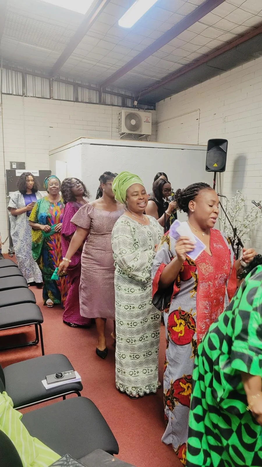 Group of women standing in a line, participating in a gathering or event, some holding papers or objects, inside a room with white brick walls, a speaker, and an air conditioning unit visible on the wall.