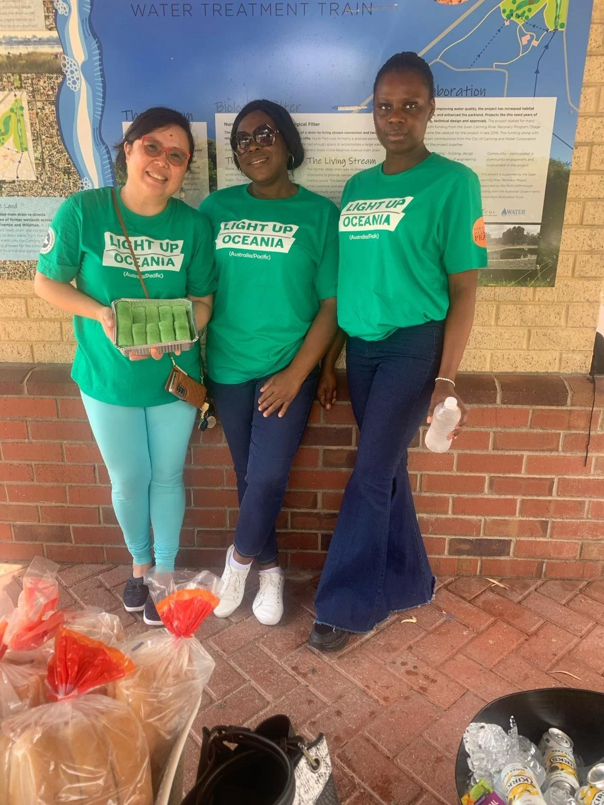 Three women standing in front of a blue information board, wearing green shirts that say "Light Up Oceania". The woman on the left is holding a tray of green treats. There are bags of bread and cans of drinks on the ground in front of them.