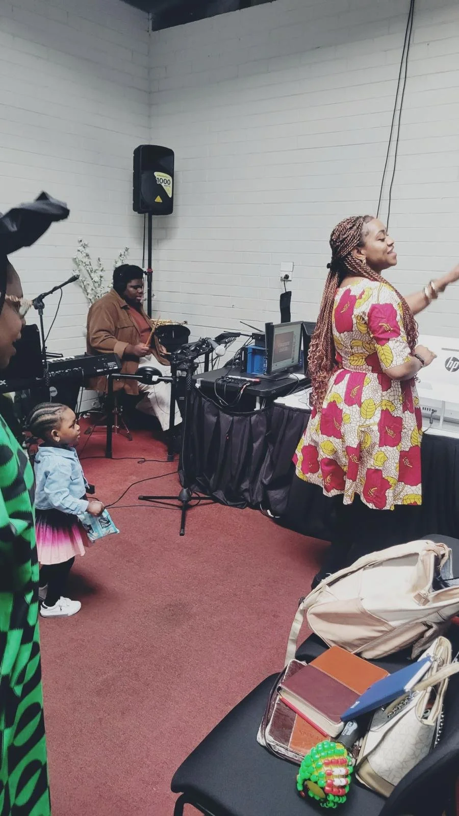 A woman in a floral dress leading a singing or speaking event in a room with a small band playing in the background, including a drummer and keyboard player. A child and a woman are watching attentively.