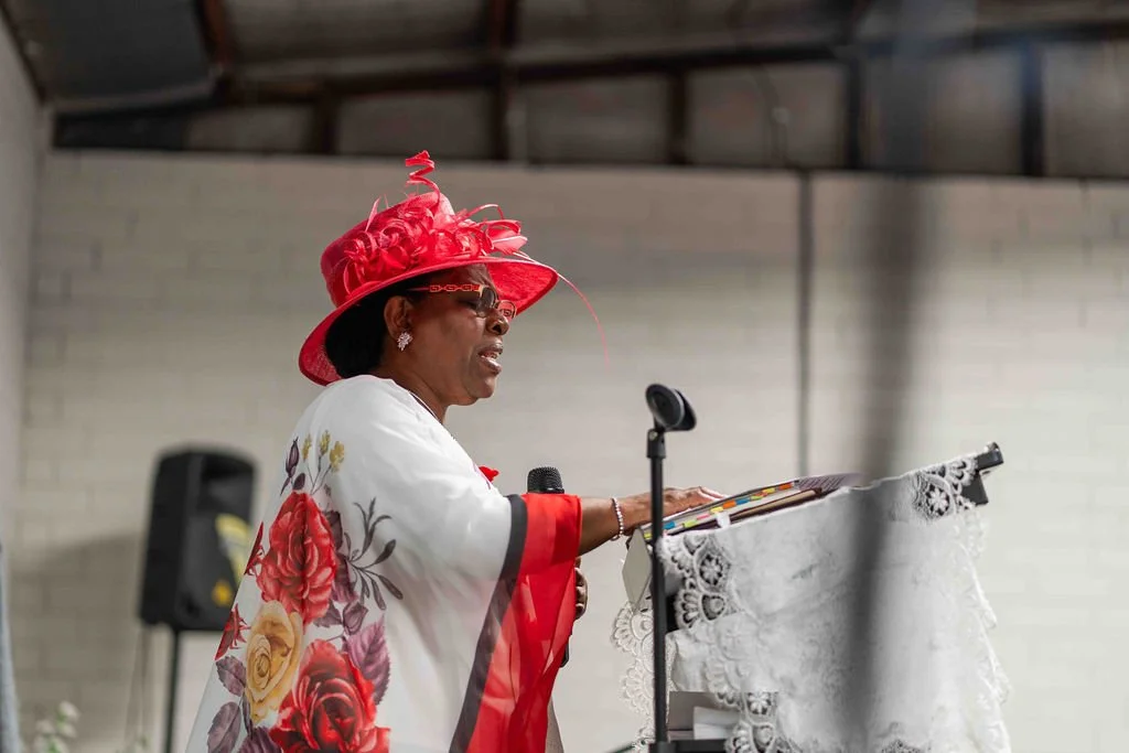 Woman in a red decorated hat speaking at a microphone behind a lace-covered podium