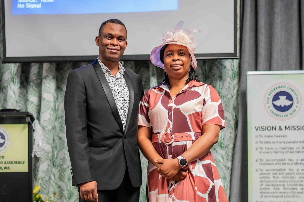 Man in a black suit and woman in a pink patterned dress and hat pose at a church event.