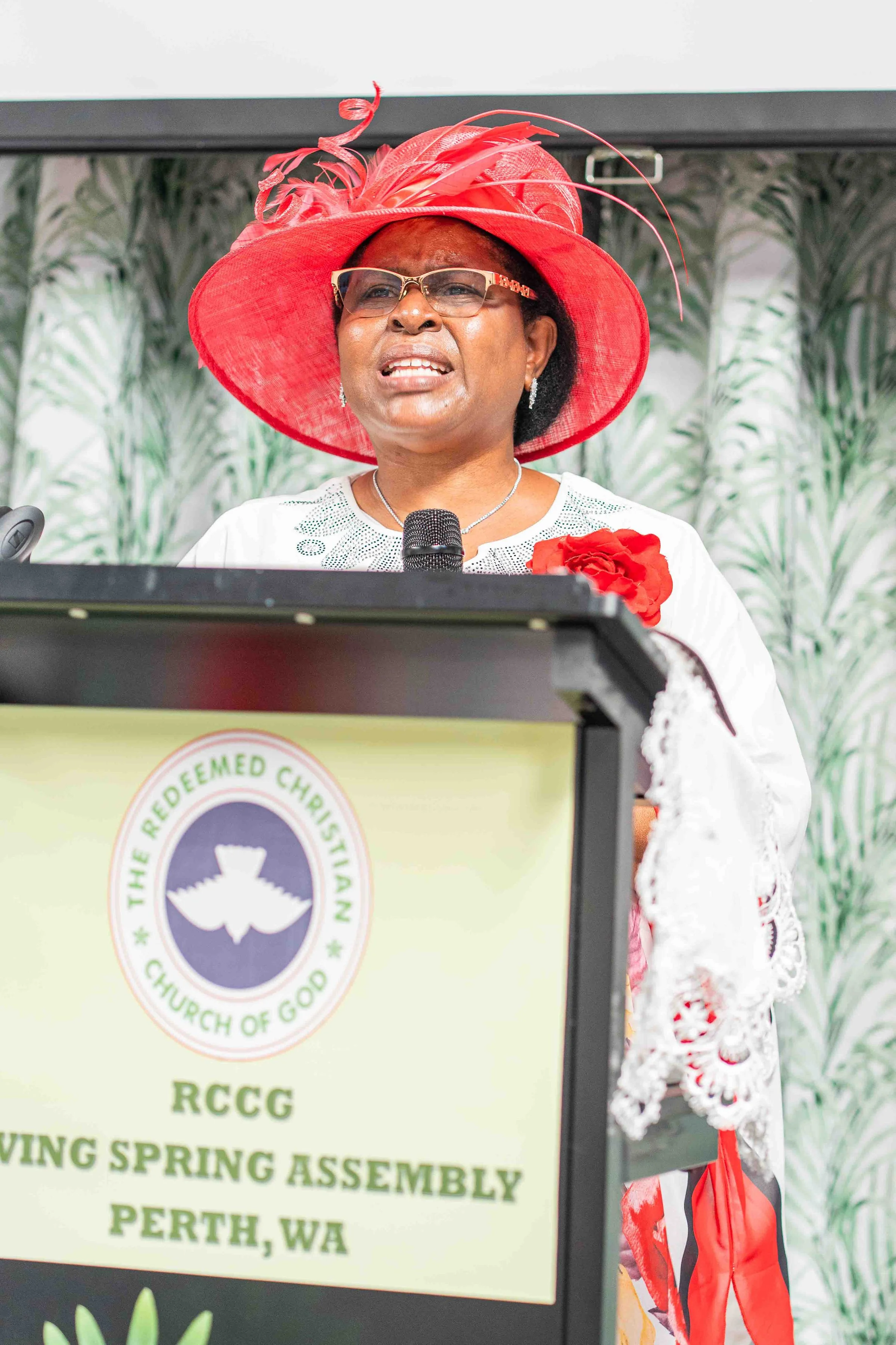 Woman in a red feathered hat and glasses speaking into a microphone at a church podium in Perth, Western Australia.