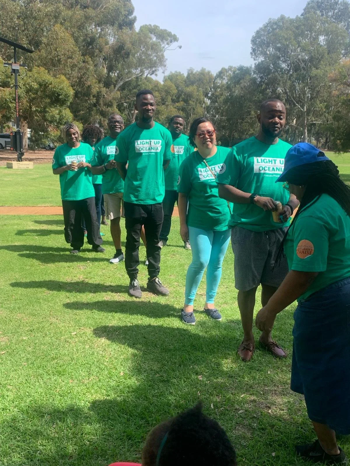 A group of people standing in a line outdoors on a grassy field, participating in a charity event called 'Light Up Oceania,' with trees and a clear sky in the background.