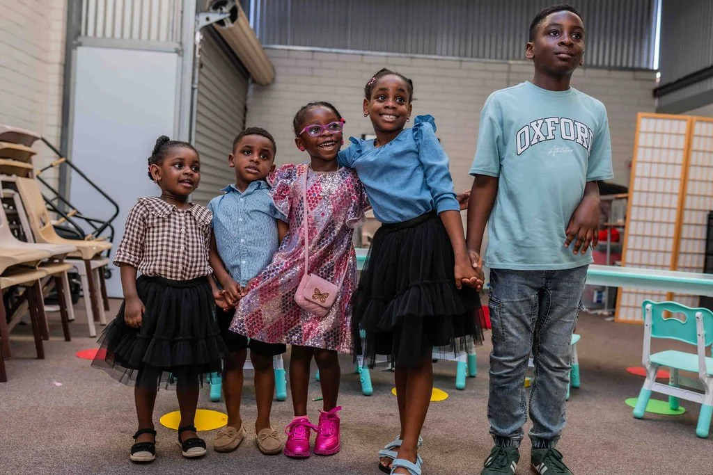 Five smiling Black children standing together in a school or community activity room.