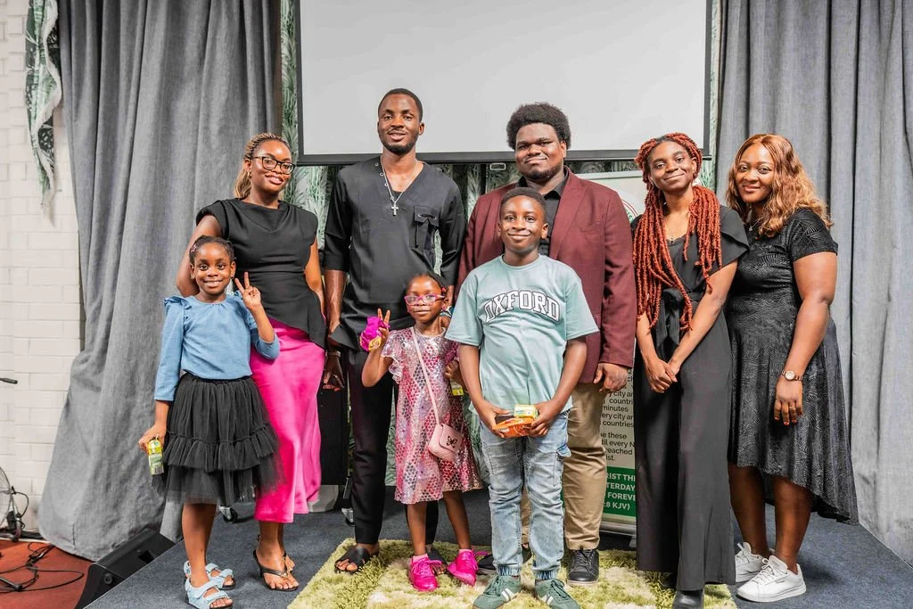Group of five adults and three children posing together on a stage in front of gray curtains and a projection screen