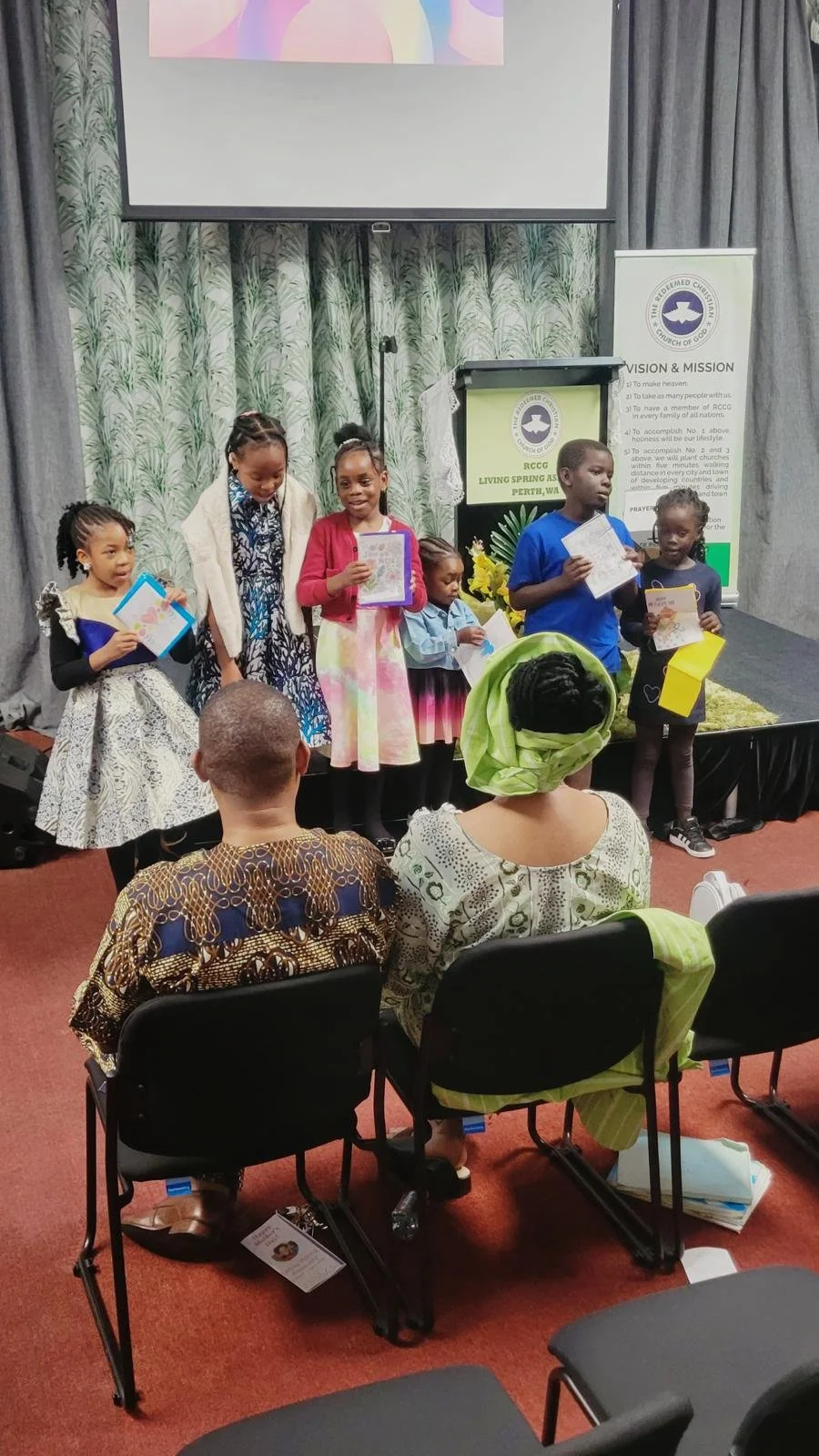 Children standing on a stage receiving awards or certificates, with two adults seated in the foreground watching.